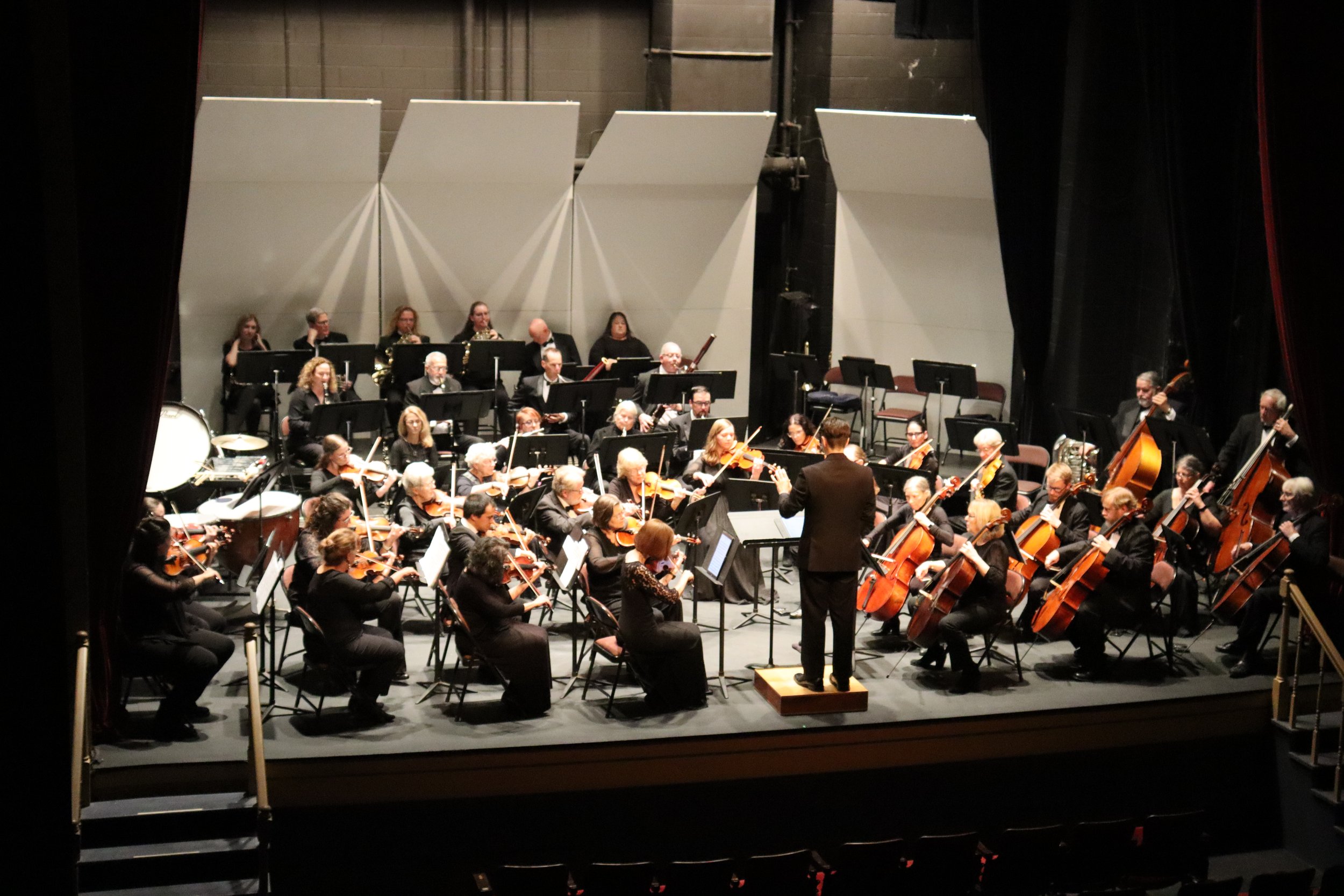 An orchestra performing on stage, viewed from backstage, with musicians playing string, brass, and percussion instruments, and a conductor leading them.