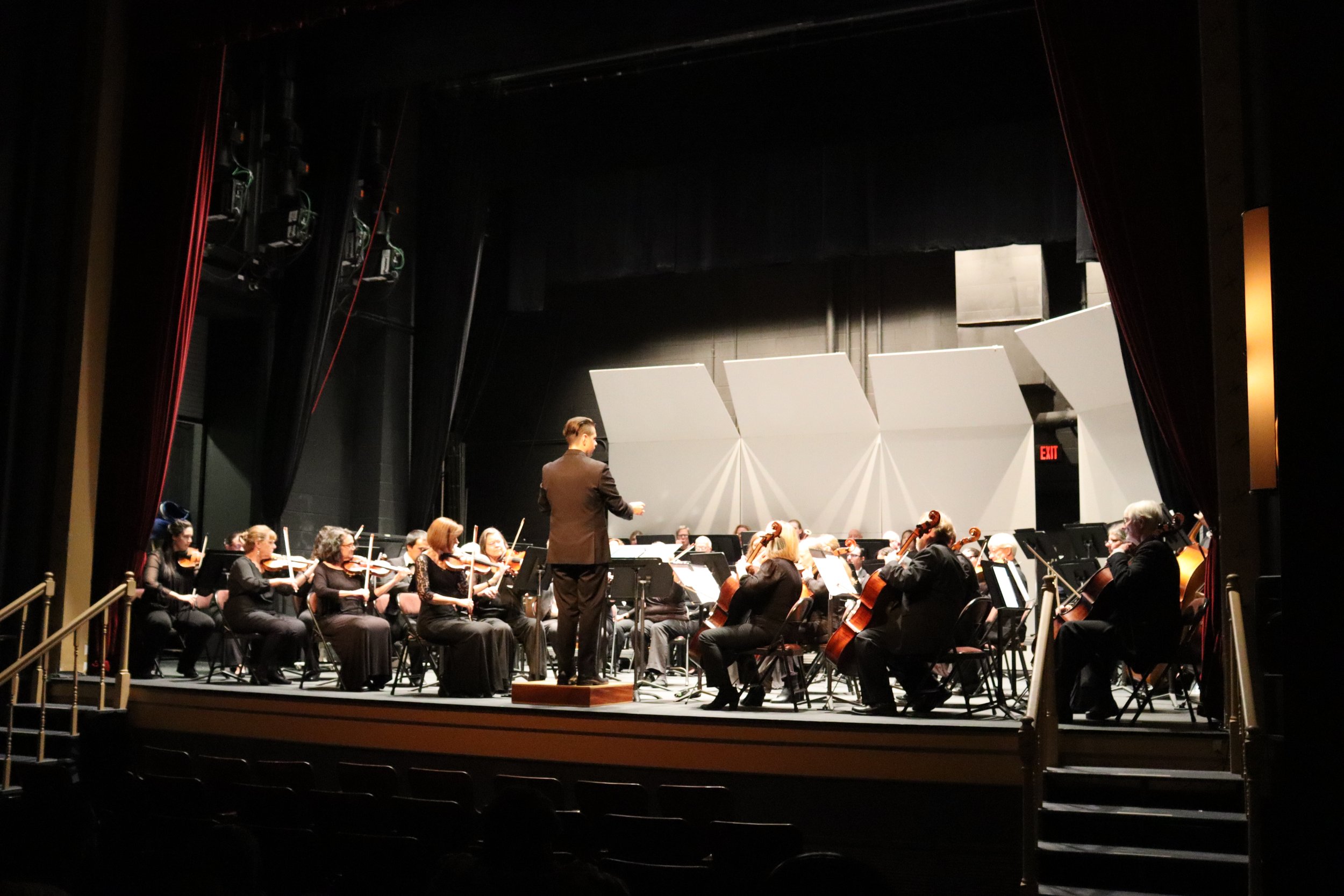 An orchestra performing on stage, with musicians seated and a conductor standing in front, in a theater with black walls and stage equipment visible.