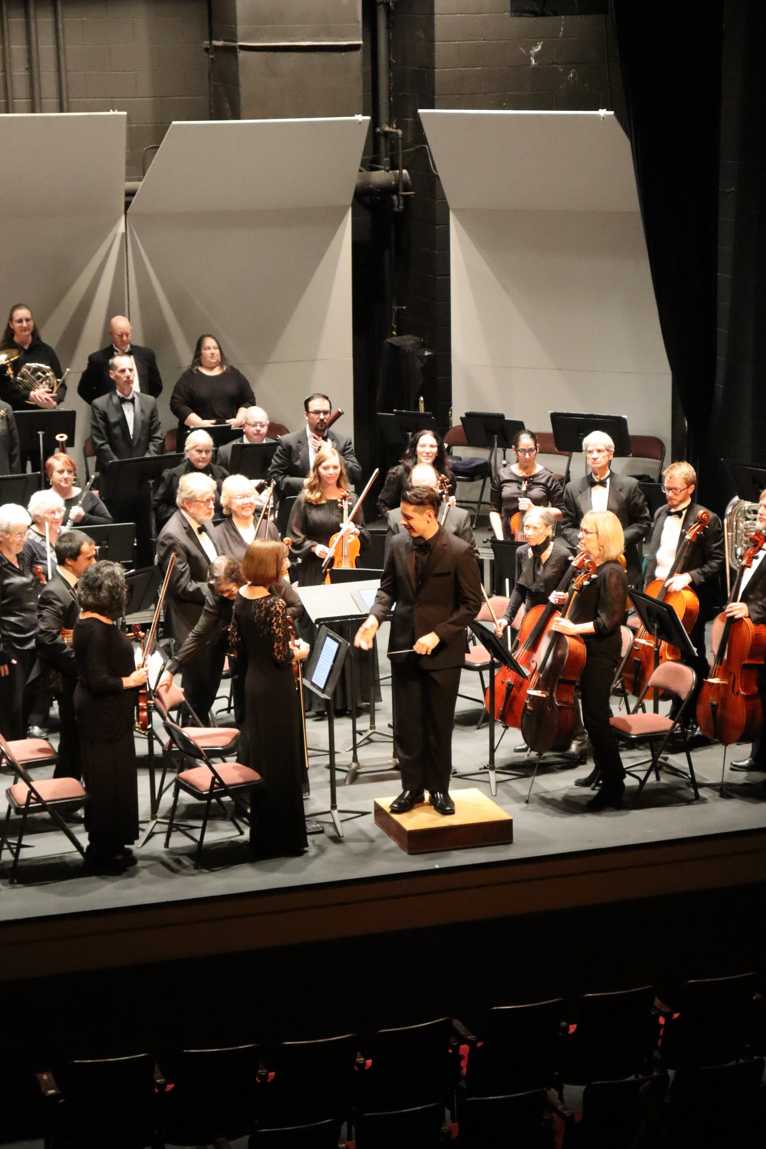 An orchestra on stage after a performance, with conductor in the center, members holding instruments, and the stage setup visible in the background.