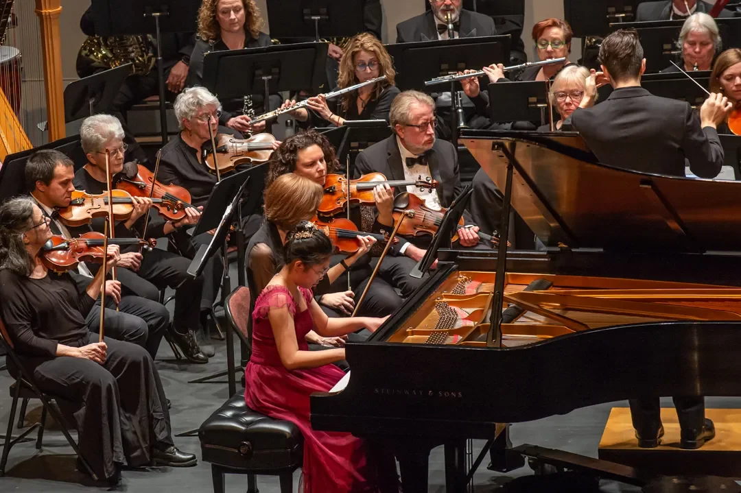 An orchestra performing on stage with a young girl playing grand piano in the foreground.