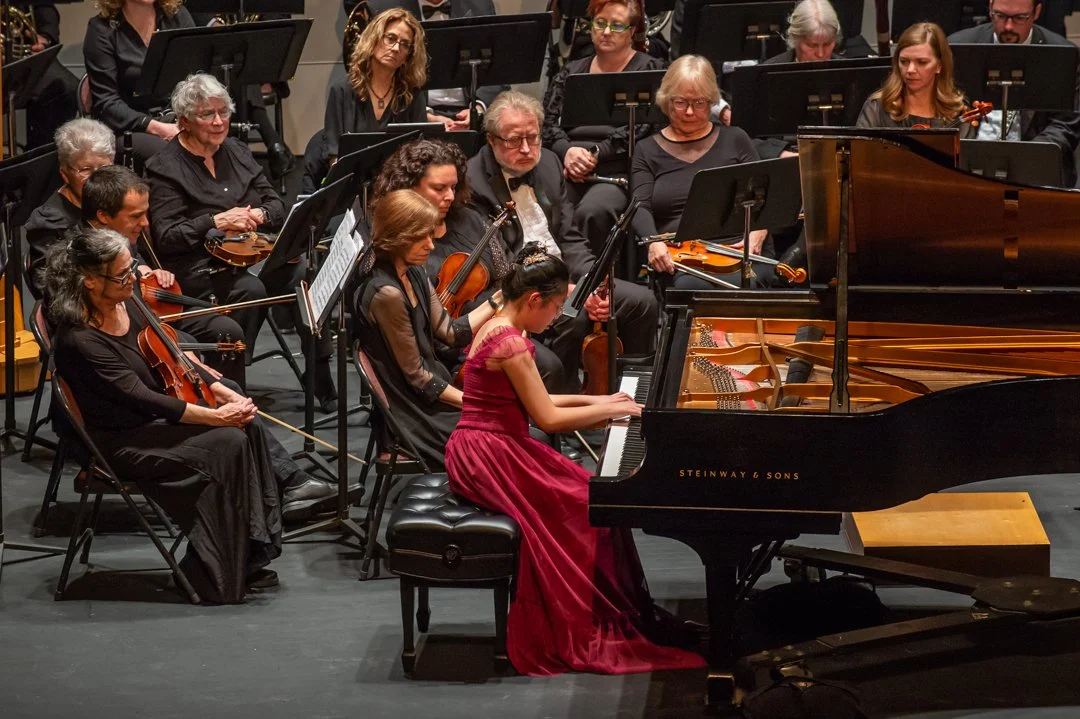 A young woman in a red gown playing a grand piano during a classical orchestra performance.