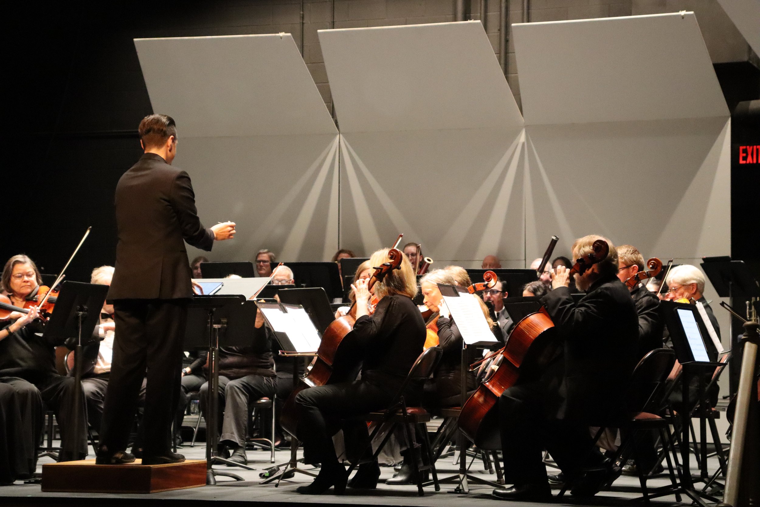 An orchestra performing on stage directed by a conductor, with musicians playing violins and cellos in front of large grey acoustic panels, with an exit sign visible on the right.