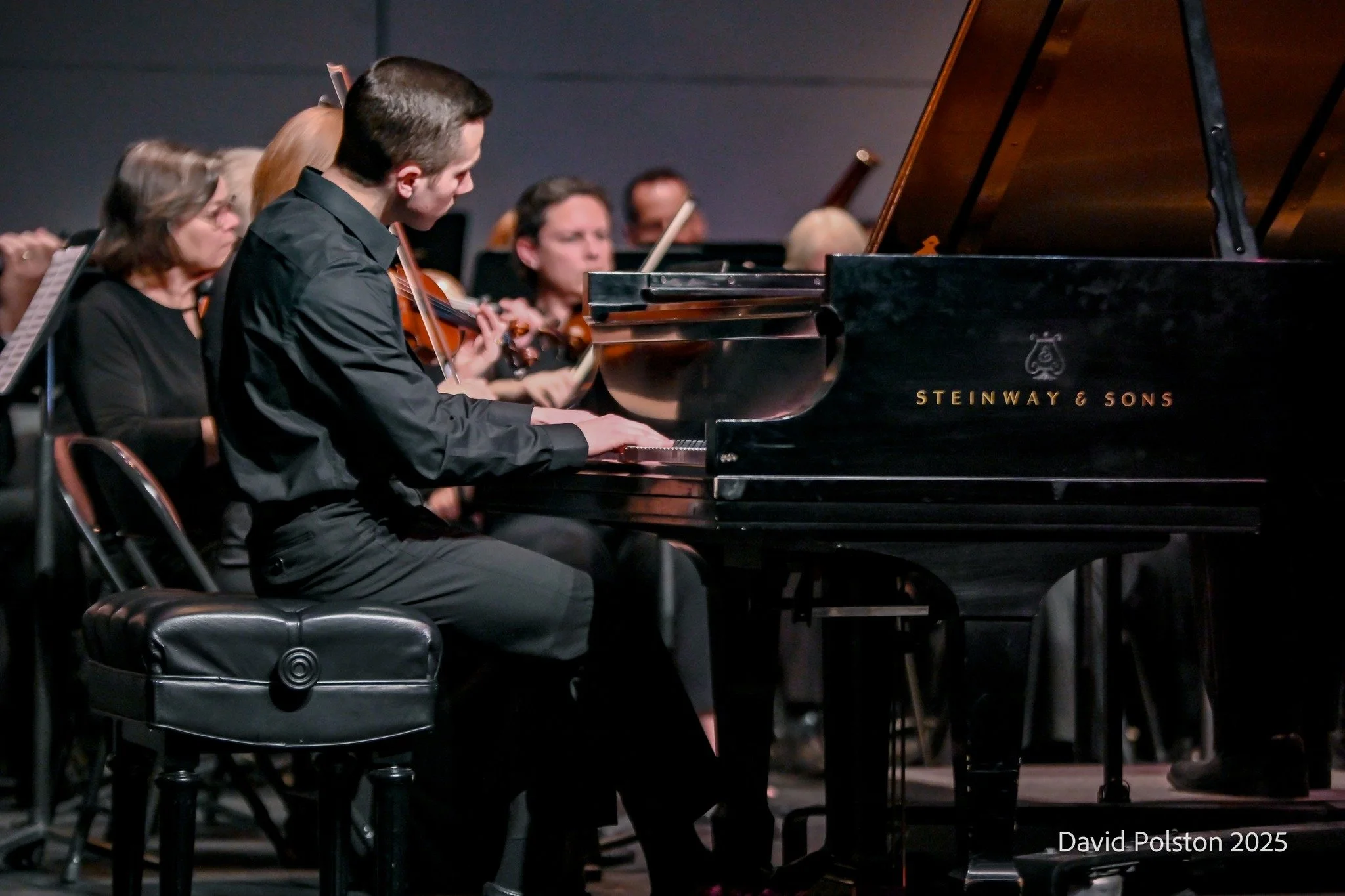 A group of musicians performing on stage, with the focus on a young man playing the piano and other musicians playing string instruments in the background.