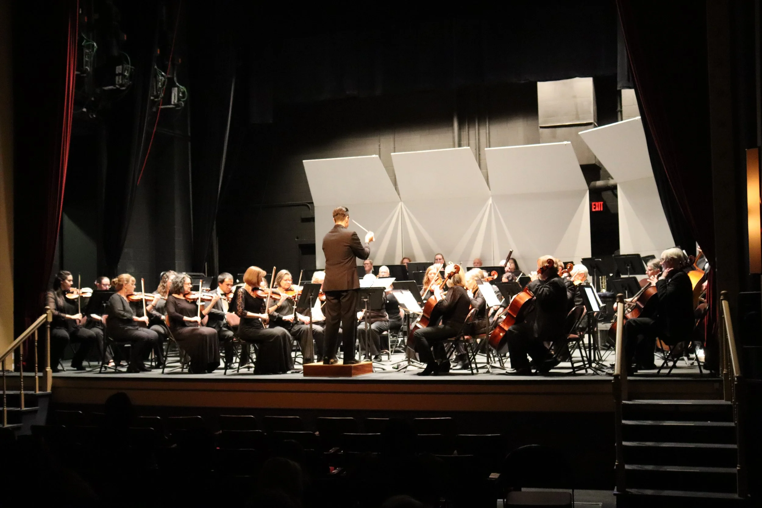 Orchestra performing on stage with conductor leading, seen from the audience perspective, with stage lights illuminating musicians and their instruments, dark background, and auditorium seats visible in the foreground.