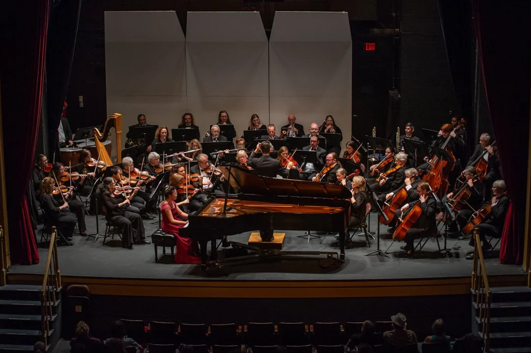 An orchestra performing on stage with a pianist in a red dress, conductor, and various musicians playing strings, woodwinds, and brass instruments, with audience seating visible in the foreground.