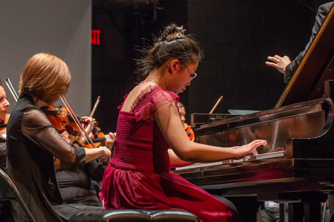 Young woman playing the piano during an orchestra performance, with violinists playing in the background.