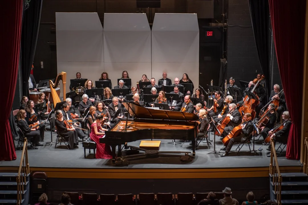 Orchestra performing on stage with a grand piano at the center, surrounded by string, wind, and percussion instruments, in a concert hall.