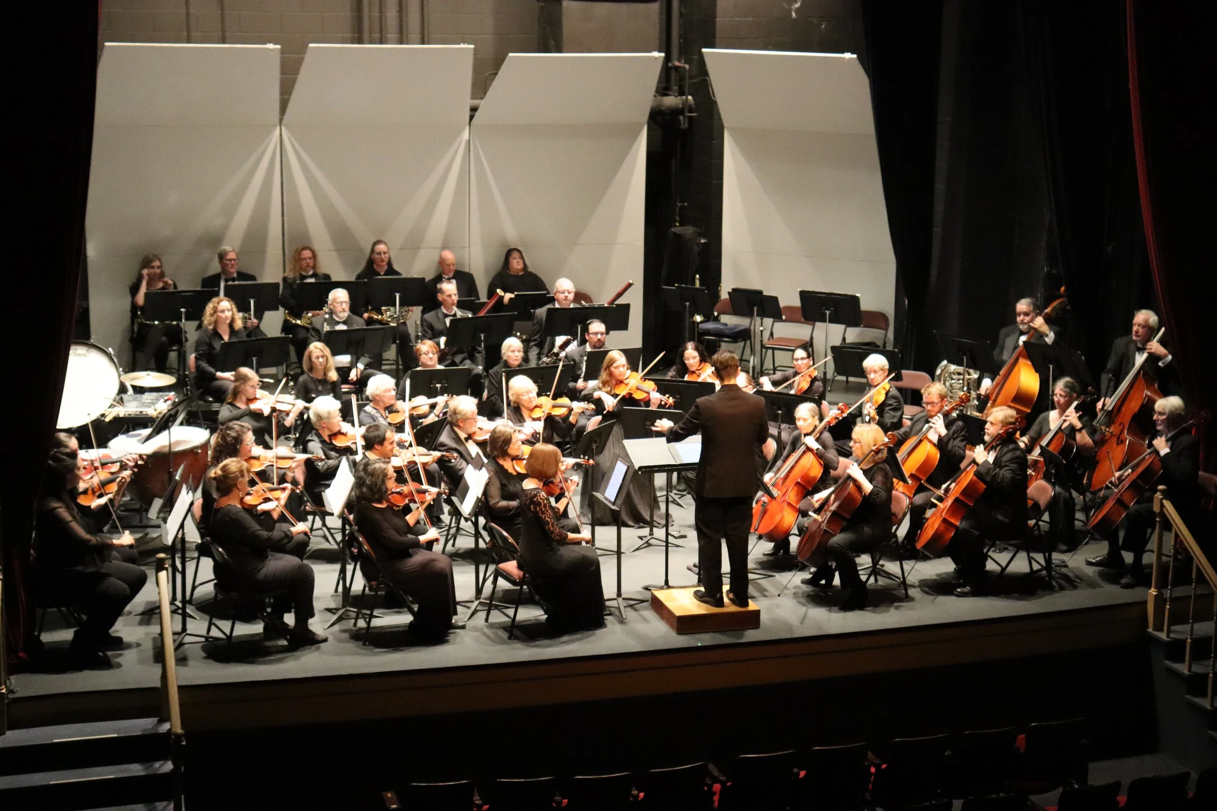 An orchestra performing on stage with musicians playing violins, cellos, and wind instruments, conducted by a conductor in front of them.