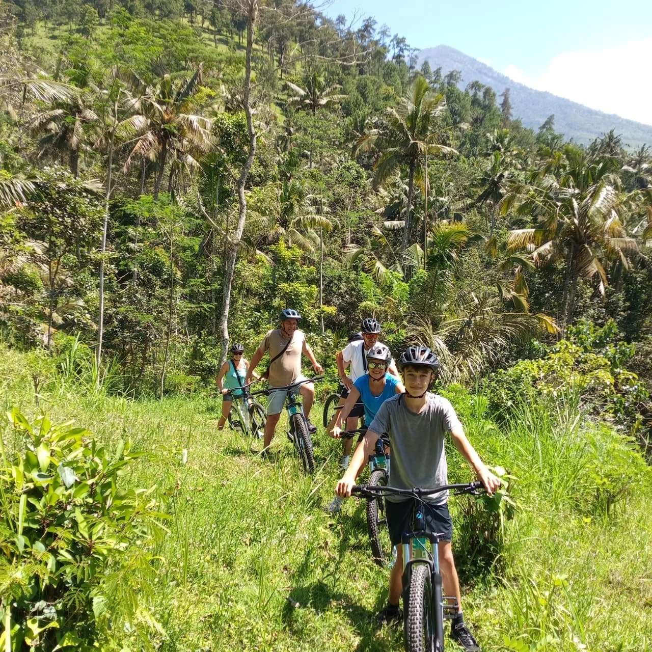 A group of people riding bicycles along a lush, green trail surrounded by tall trees and dense foliage, with mountains visible in the background on a sunny day.
