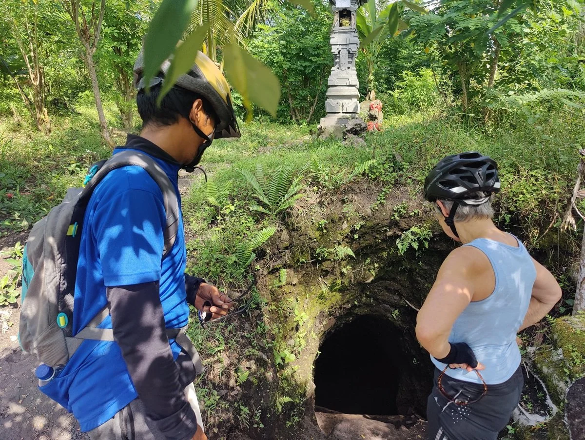 Two cyclists, a man and a woman, wearing helmets and cycling gear, look into a dark cave opening in a lush, green forest.