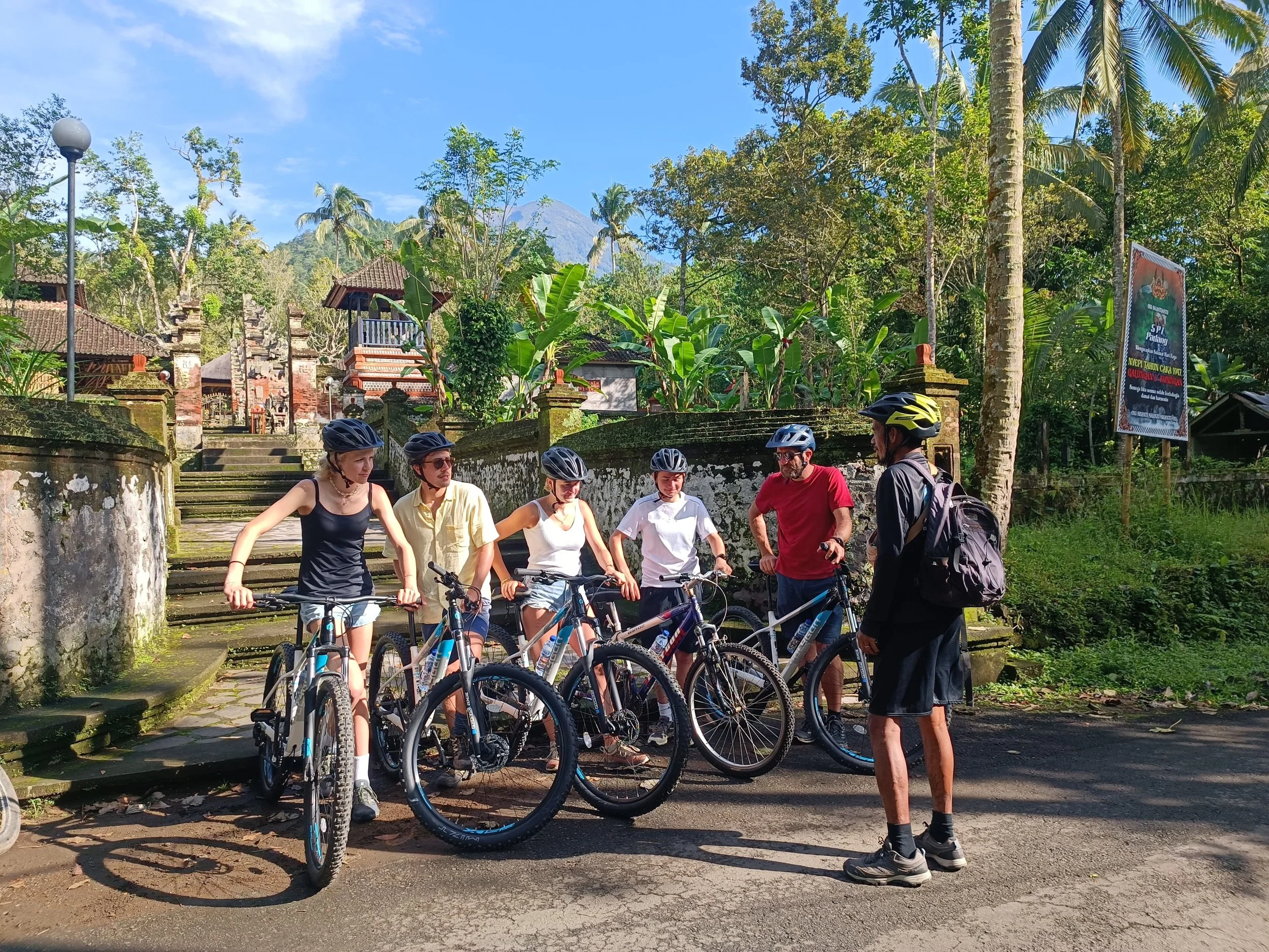 Group of six friends with mountain bikes and helmets, talking to a man with a backpack, in a tropical setting with trees, stairs, and traditional buildings in the background.
