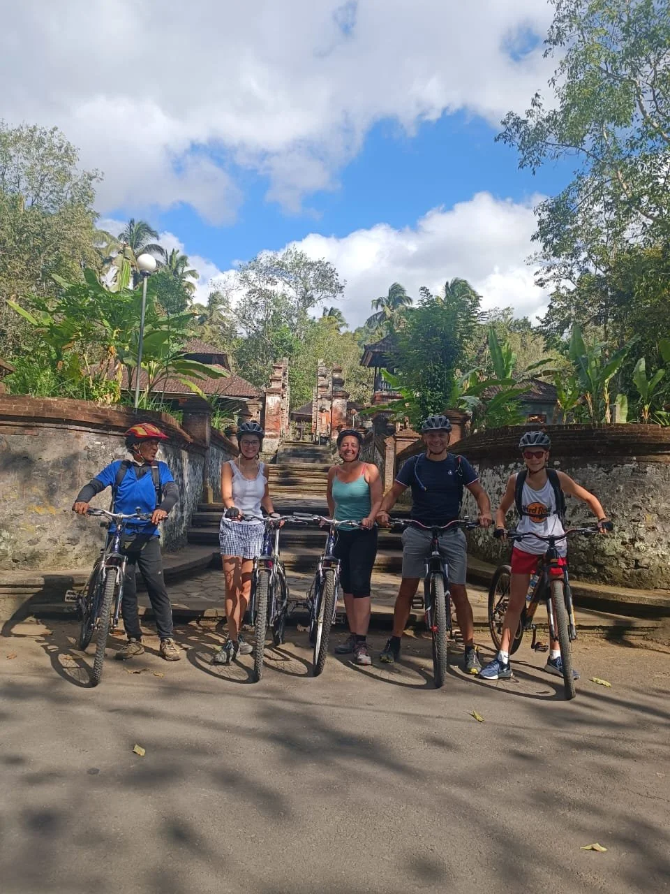 Group of five people with bicycles standing on a paved path outdoors, wearing helmets, with trees and buildings in the background, under partly cloudy sky.