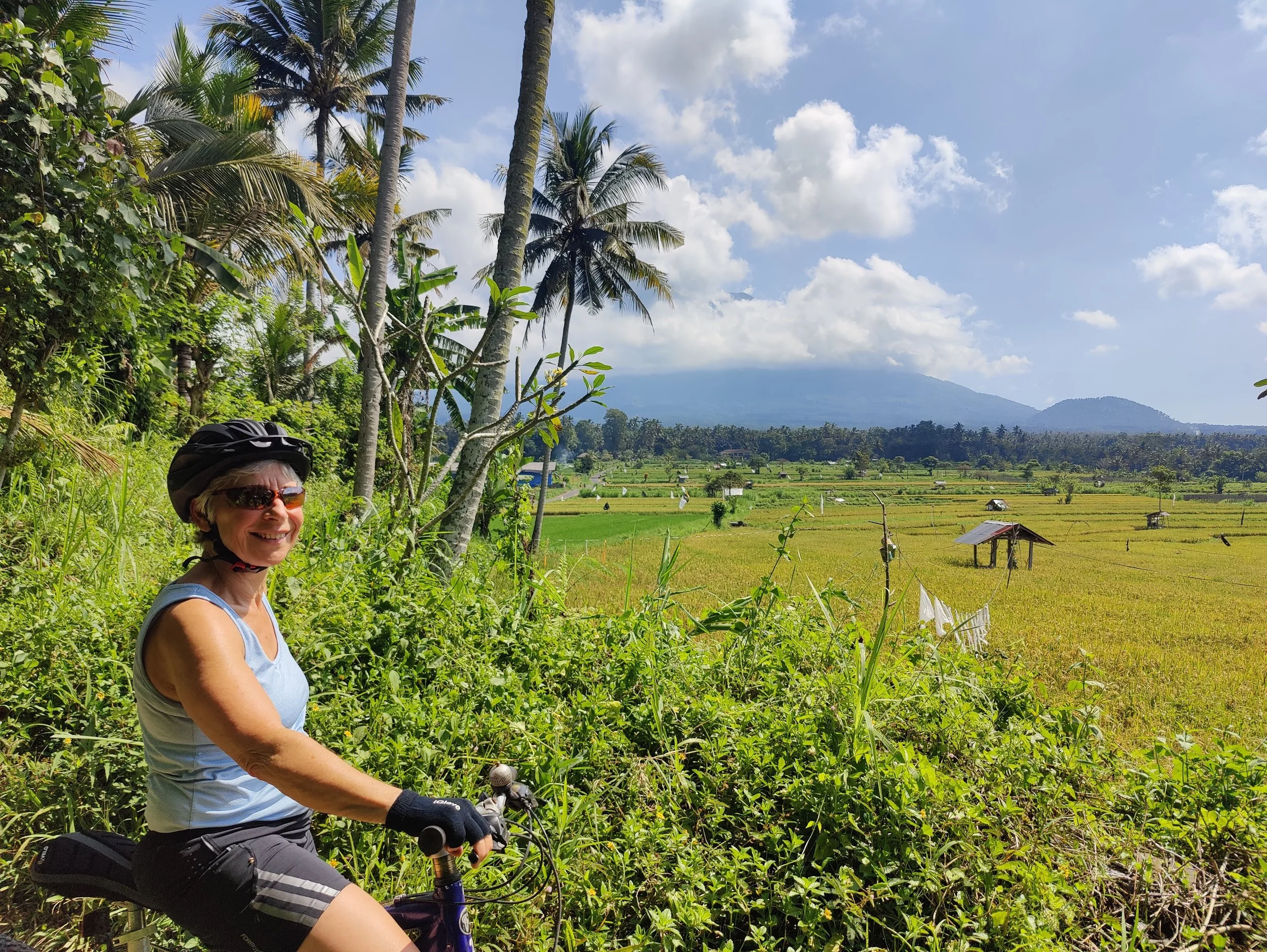 A woman in cycling gear, wearing sunglasses and a helmet, smiling while riding a bicycle on a lush green path near rice fields, with palm trees and mountains in the background under a partly cloudy sky.