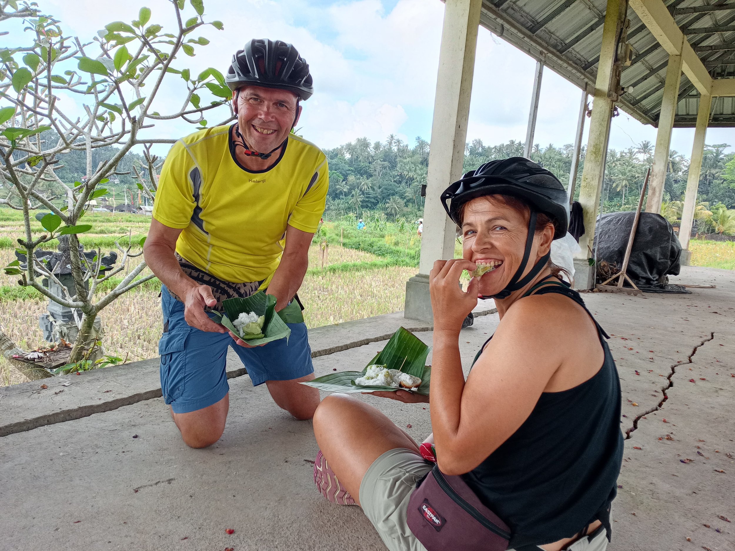 A man and woman, both wearing bicycle helmets, are smiling and enjoying traditional food wrapped in banana leaves. The man is kneeling while the woman is sitting outdoors with a lush green landscape in the background.