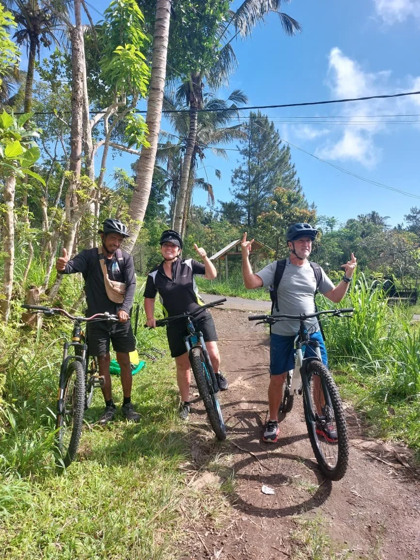 Three people in cycling gear with helmets, standing with their bikes on a dirt path surrounded by green trees and plants under a blue sky.