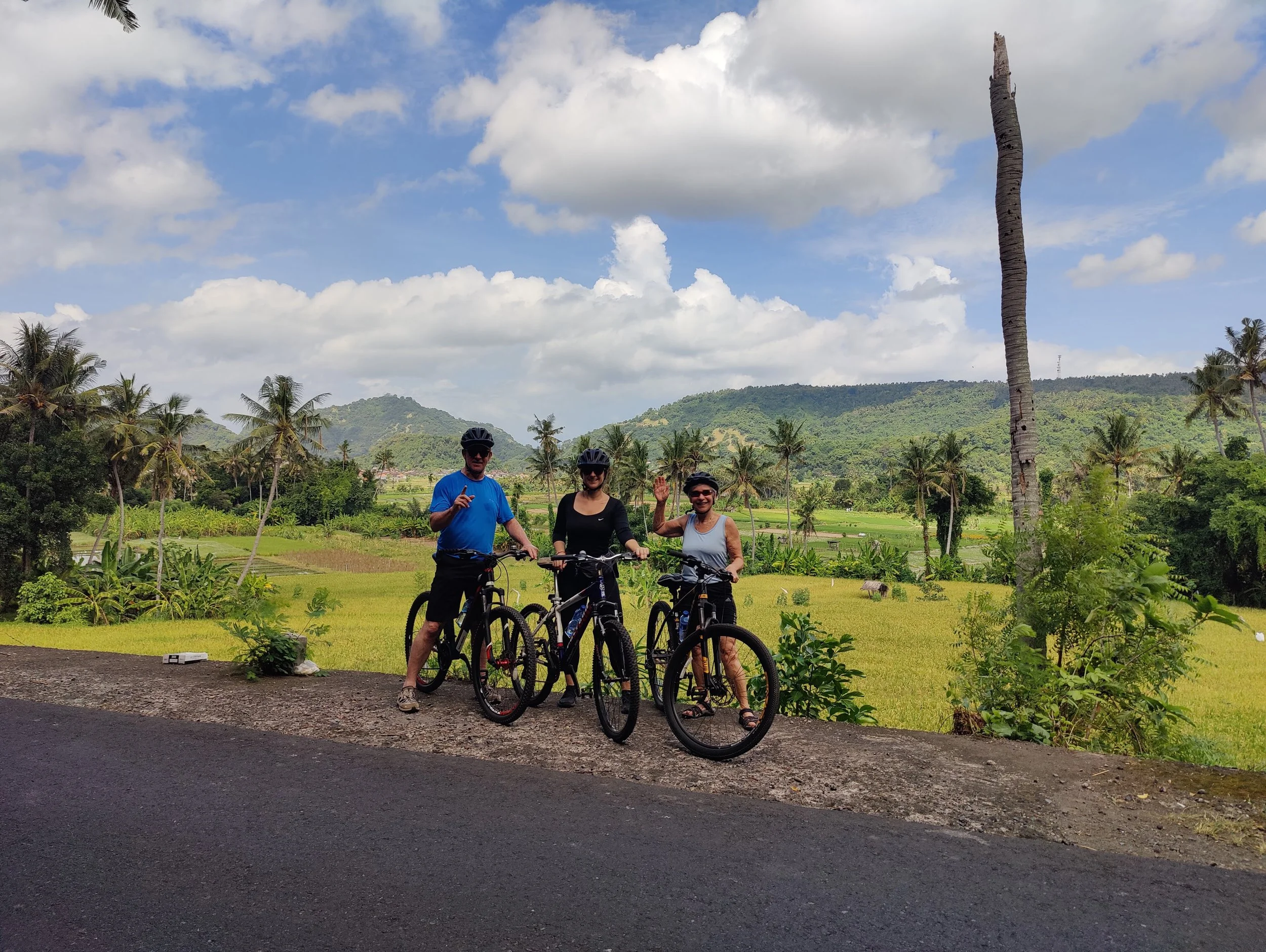 Three people in cycling gear and helmets with mountain bikes standing on the side of a road, surrounded by lush greenery and palm trees, with mountains and a partly cloudy sky in the background.