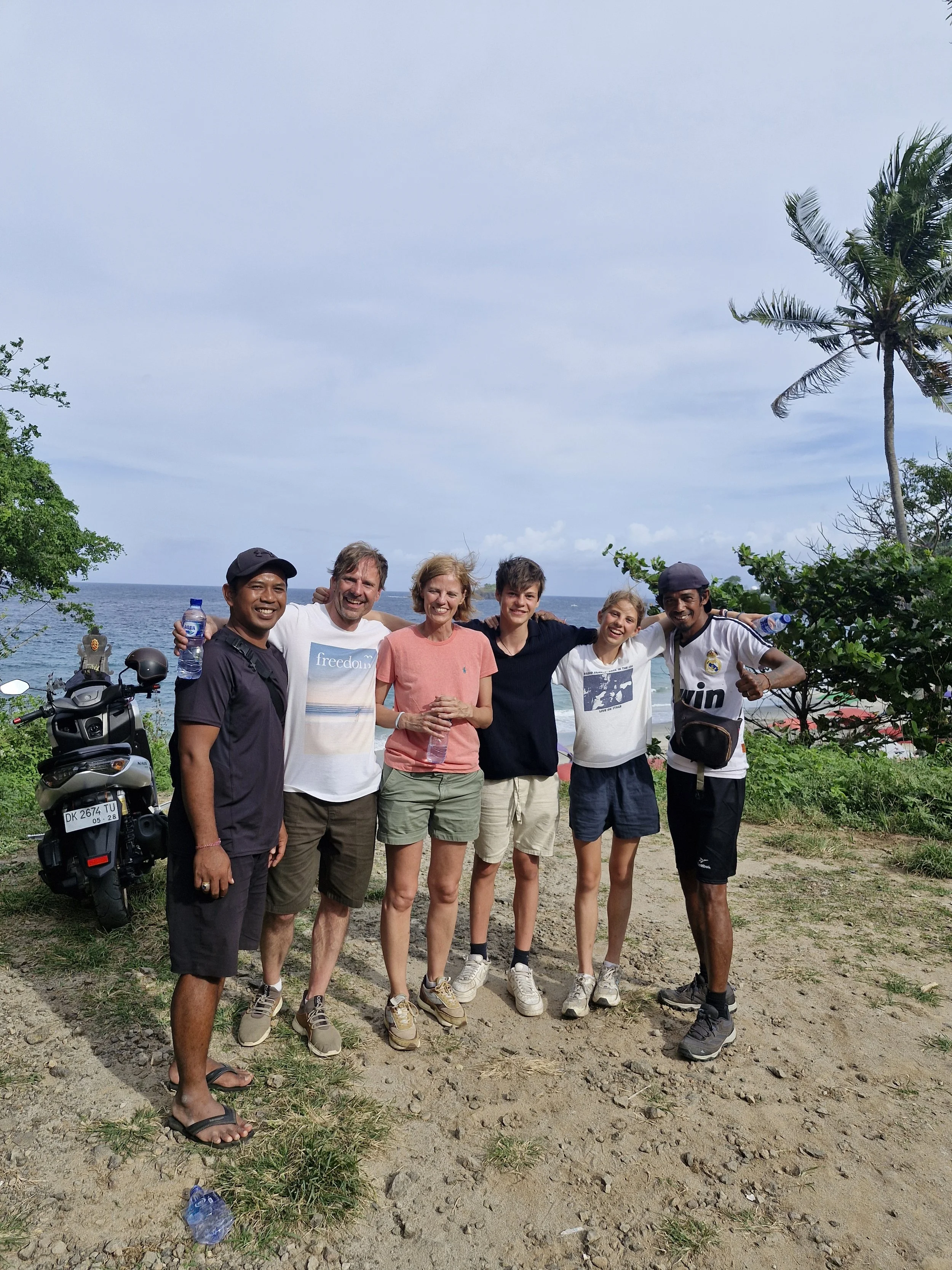 Six people standing together on a beach, smiling, with the ocean and sky in the background. There are trees and a motorcycle nearby.