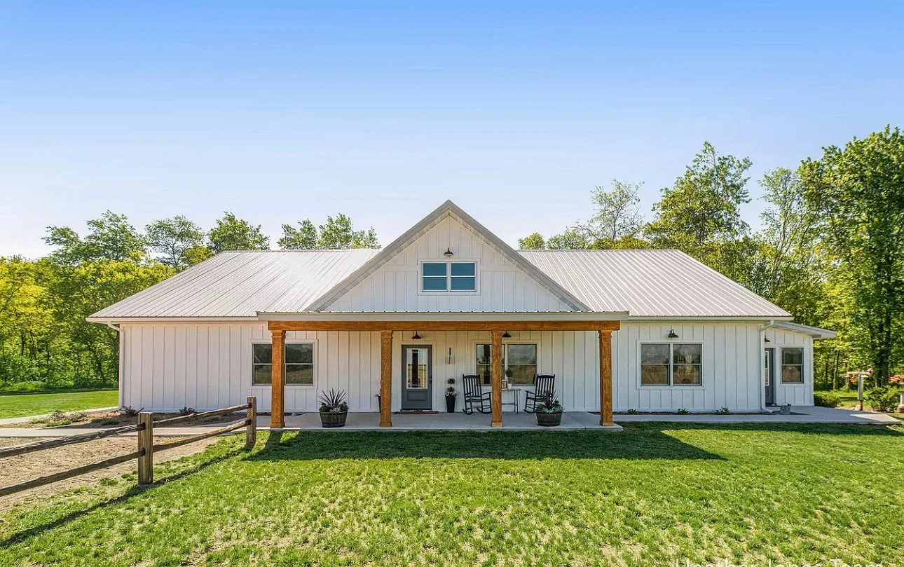 White barn-style house with a metal roof, front porch with wooden posts, and outdoor seating, surrounded by green grass and trees under a clear blue sky.