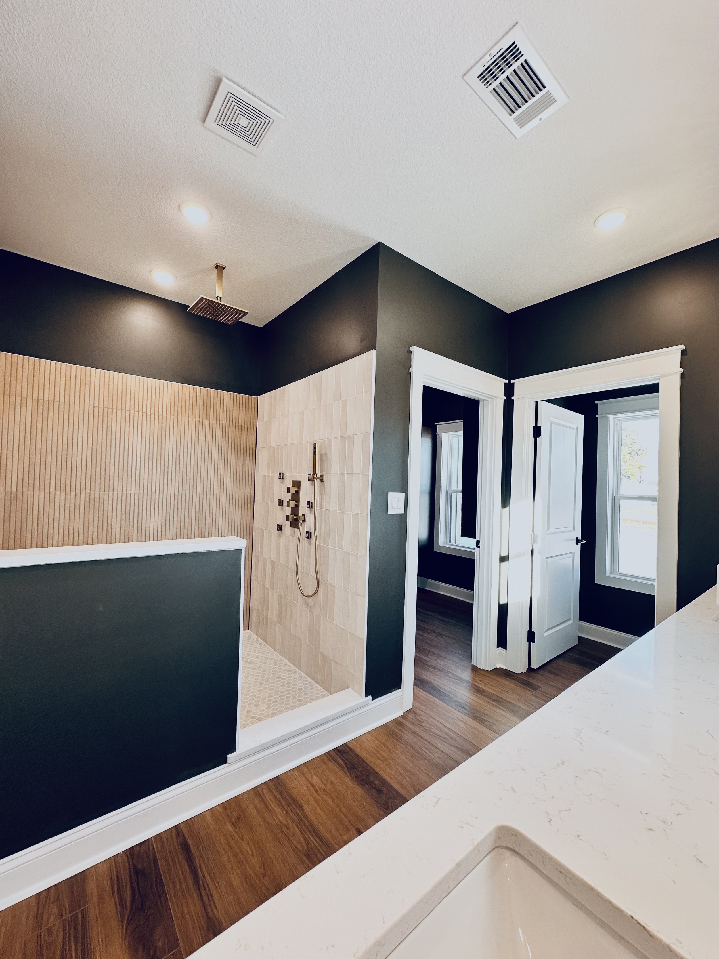 A bathroom with a walk-in shower, dark green walls, a white door, and wooden flooring seen from a white countertop.