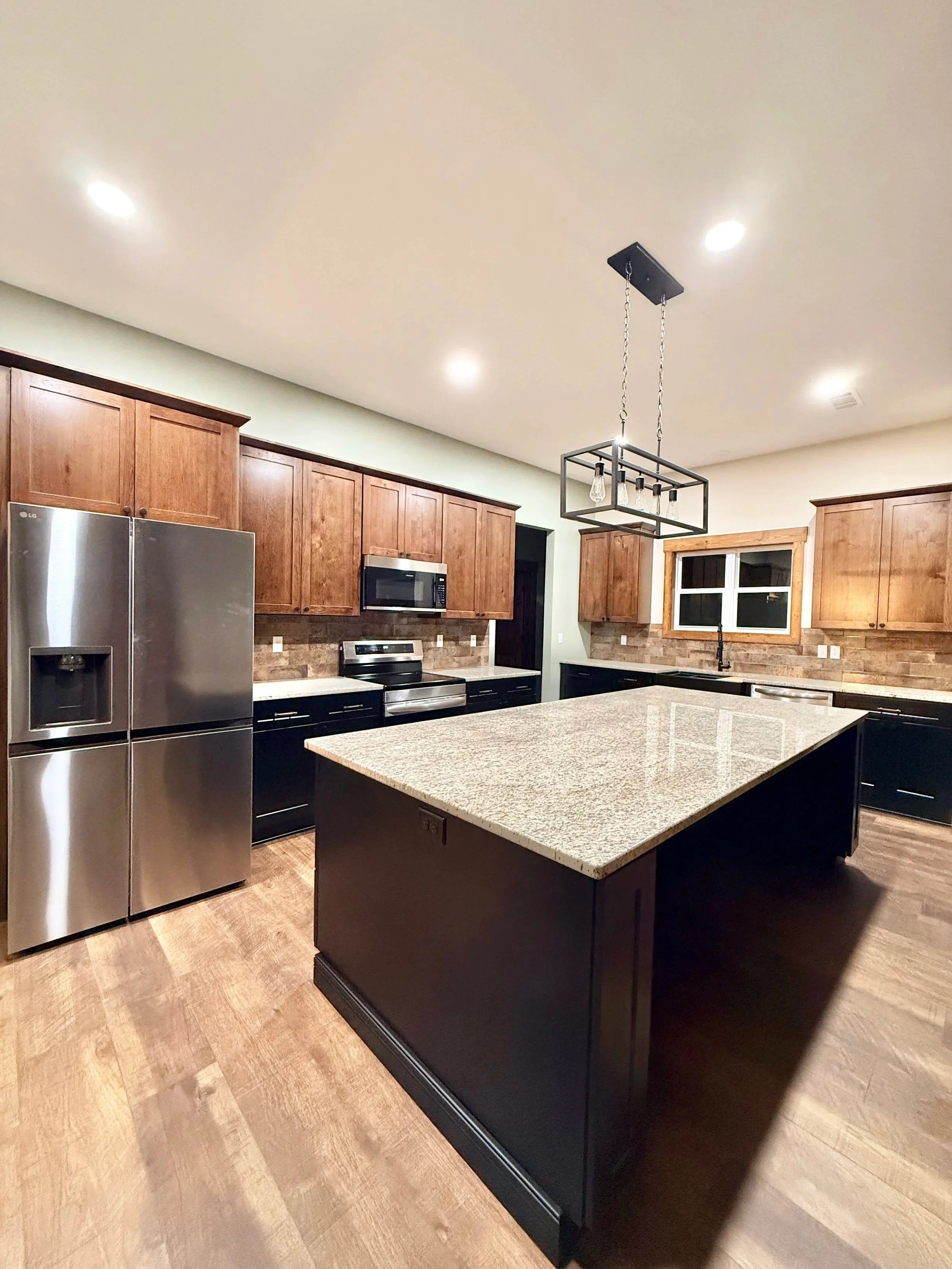 A modern kitchen featuring wooden cabinets, a stainless steel refrigerator, a microwave, a black oven, a granite island, and a geometric metal pendant light fixture.