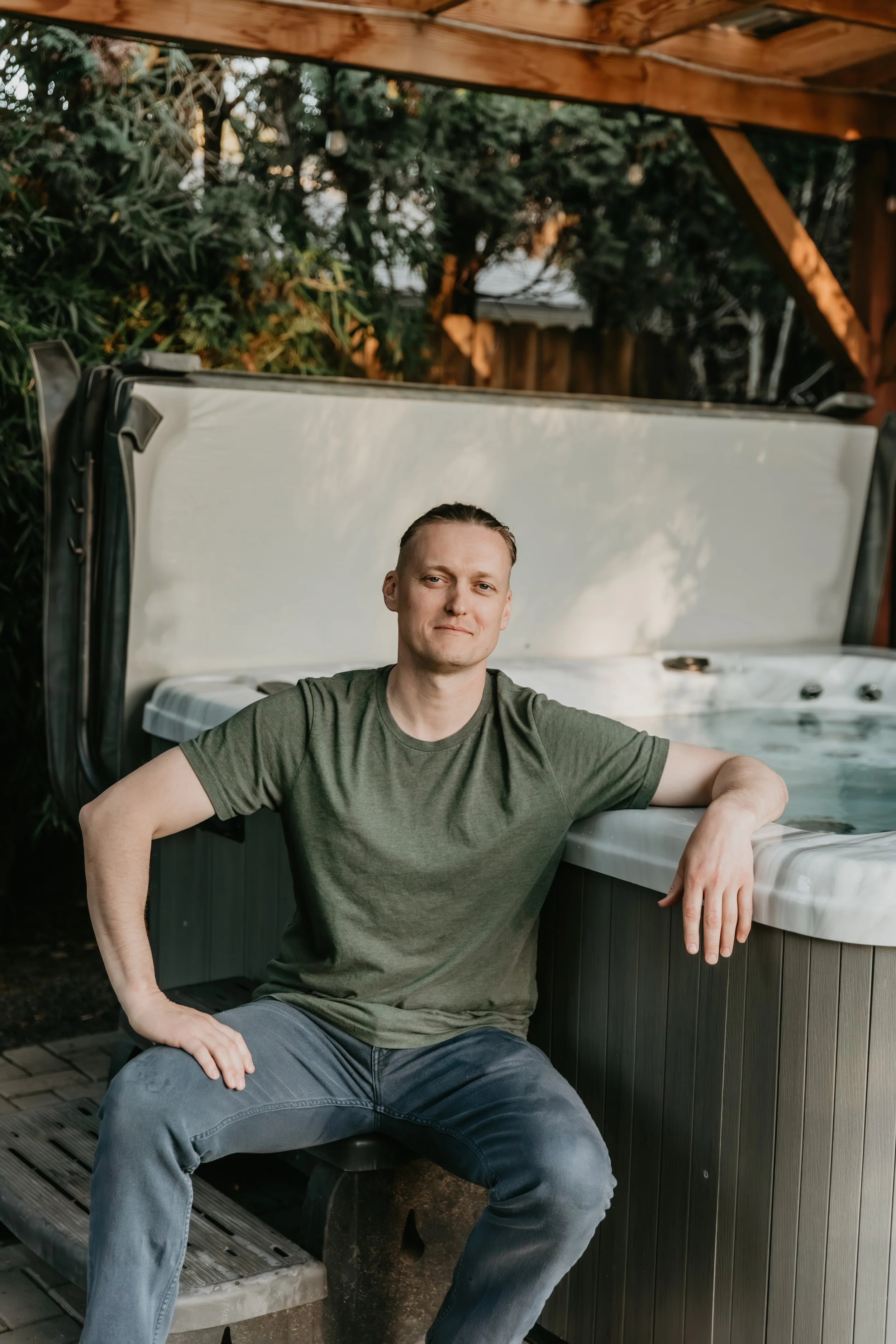 A man sitting next to an outdoor hot tub under a wooden pergola, with greenery in the background.