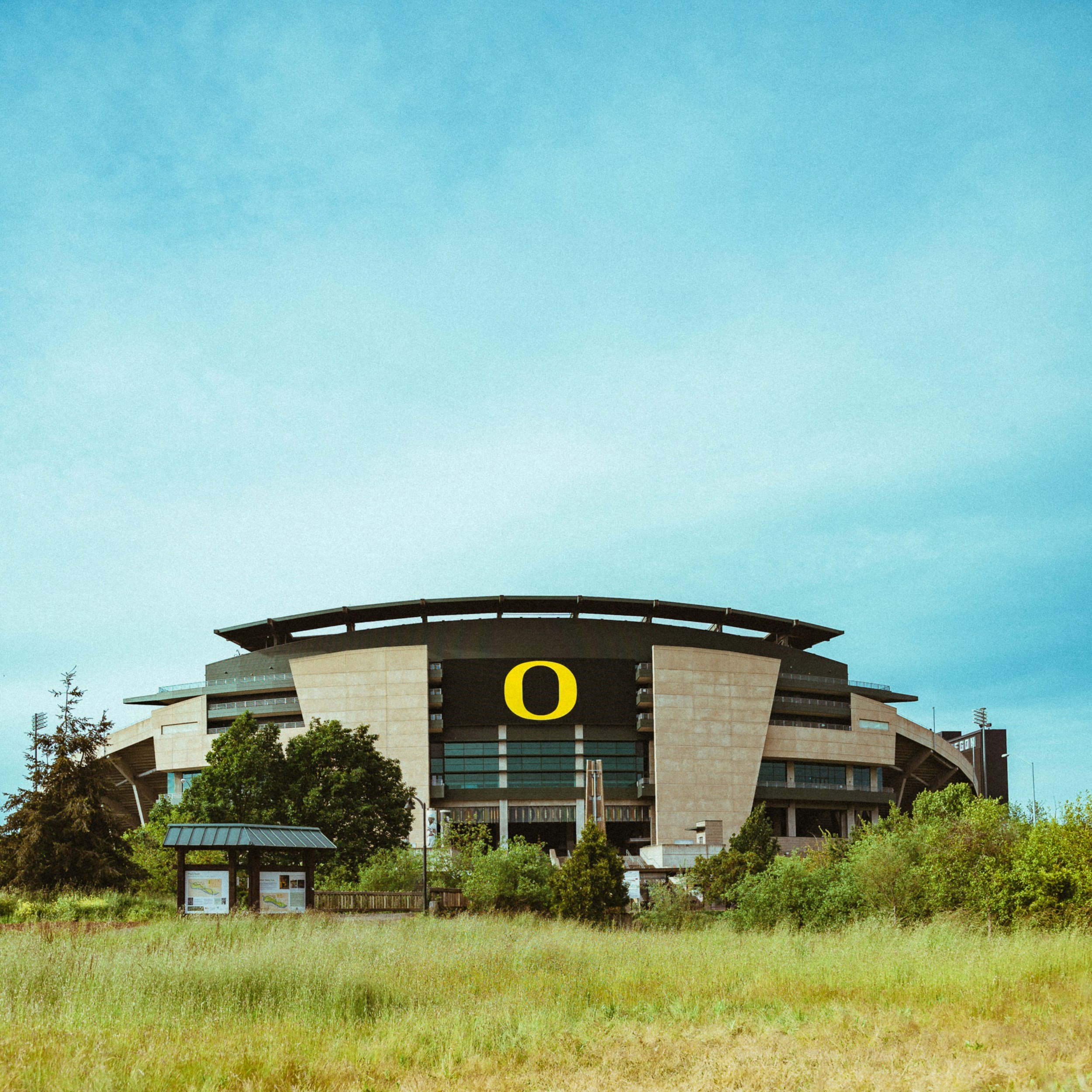 Exterior view of Autzen Stadium in Eugene, Oregon, with a large yellow 'O' sign on the front. Green grass and trees are in the foreground, and a blue sky with some clouds is above.