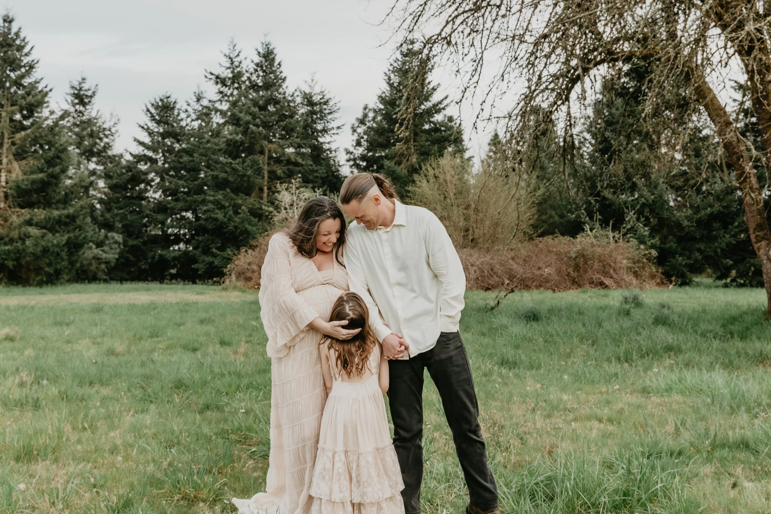 A family of three standing on a grassy field, holding hands, and smiling while looking at each other. The mother has dark wavy hair and is wearing a long beige dress, the father has long hair tied back and is wearing a white shirt and black pants, and the little girl has curly hair and is wearing a beige dress.