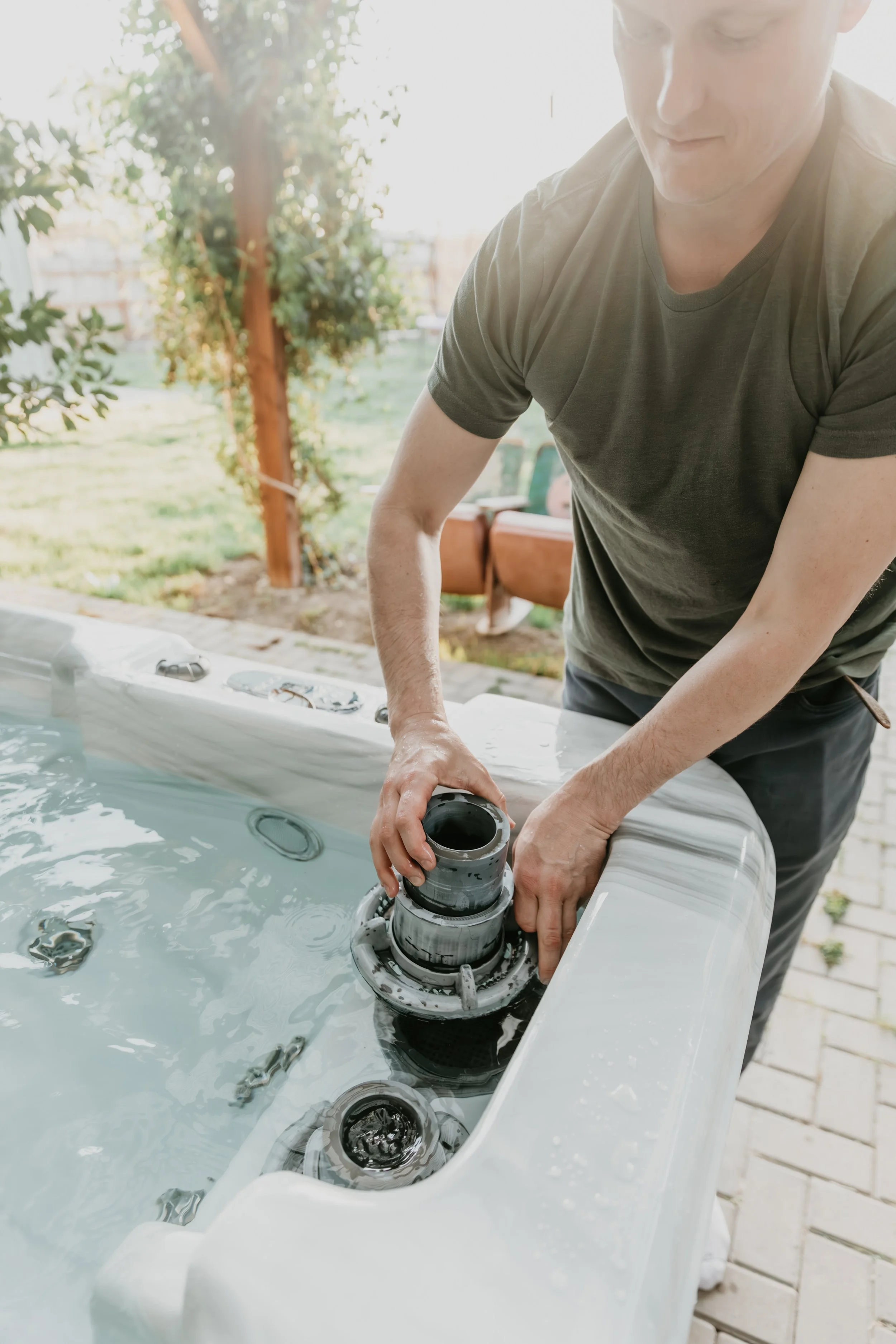 A person in a dark green t-shirt working on the jet mechanism of an outdoor hot tub or spa, with the lid removed. The hot tub is filled with water, and the scene is set in a backyard with a brick patio and trees in the background.