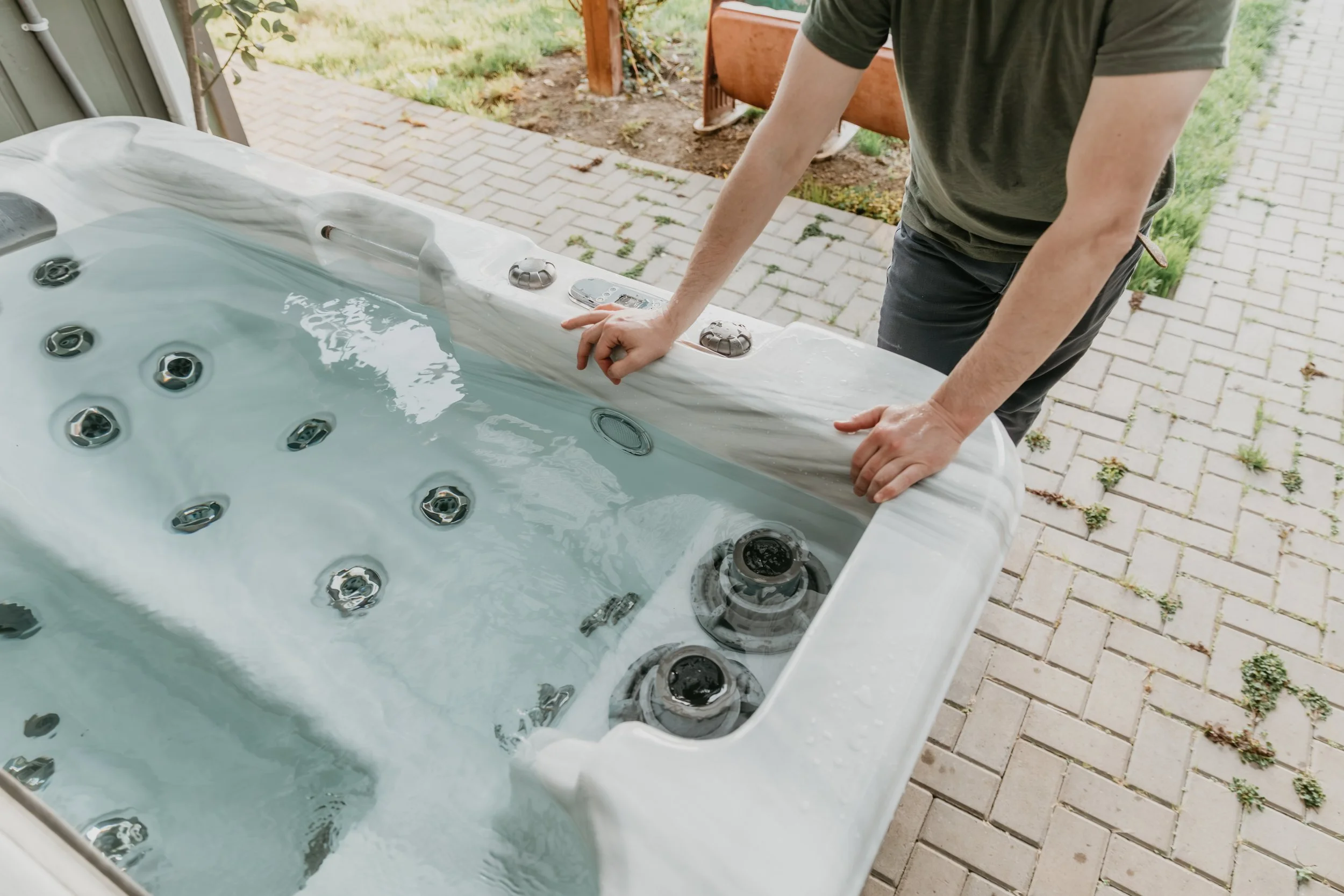 A person is leaning over the edge of a hot tub or jacuzzi outdoors, adjusting or examining the control panel. The hot tub has multiple jets and is situated on a paved patio with greenery in the background.