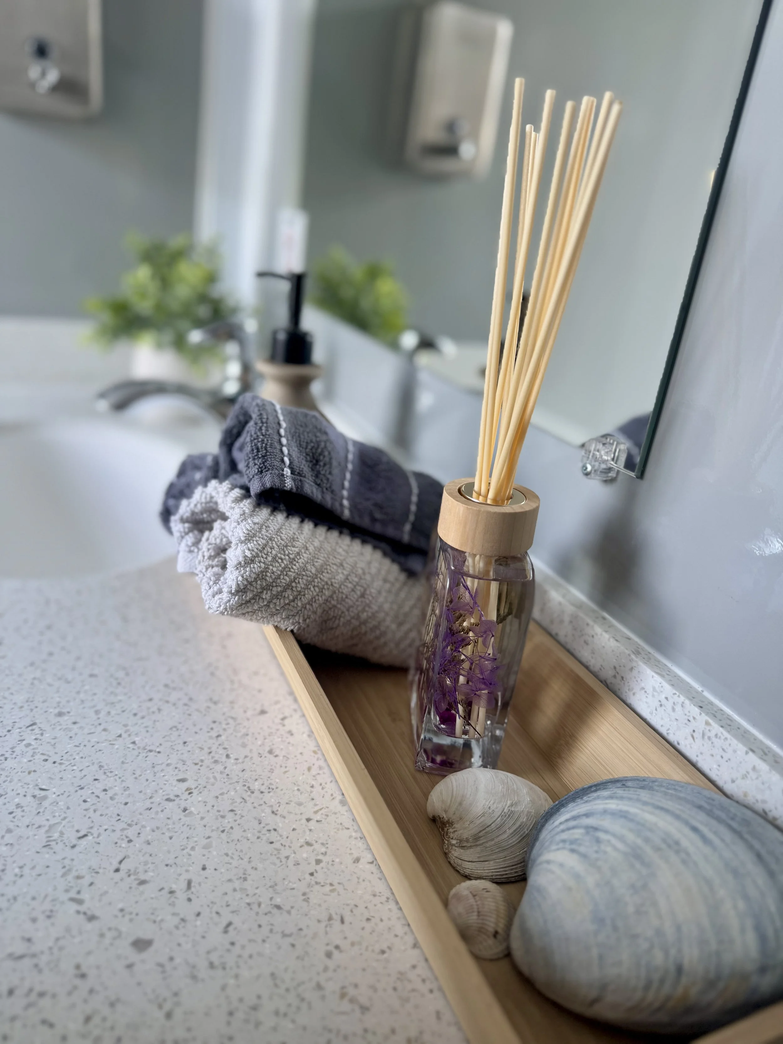 Bathroom countertop with a wooden tray holding seashells, a glass diffuser with reed sticks, and towels, with a sink and faucet in the background.