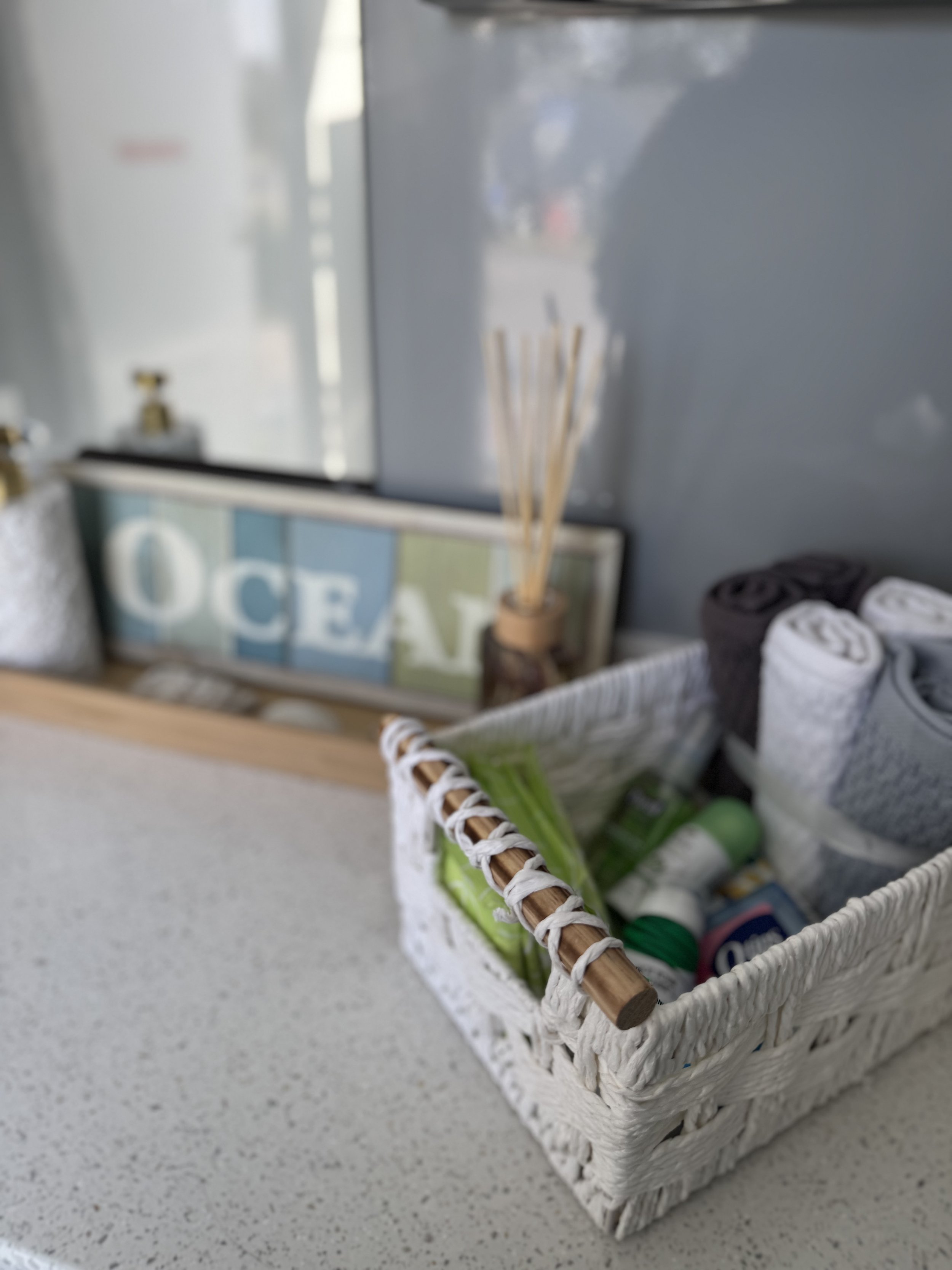 A white woven basket filled with toiletries on a light-colored countertop, with a framed decorative sign and reed diffuser in the background.