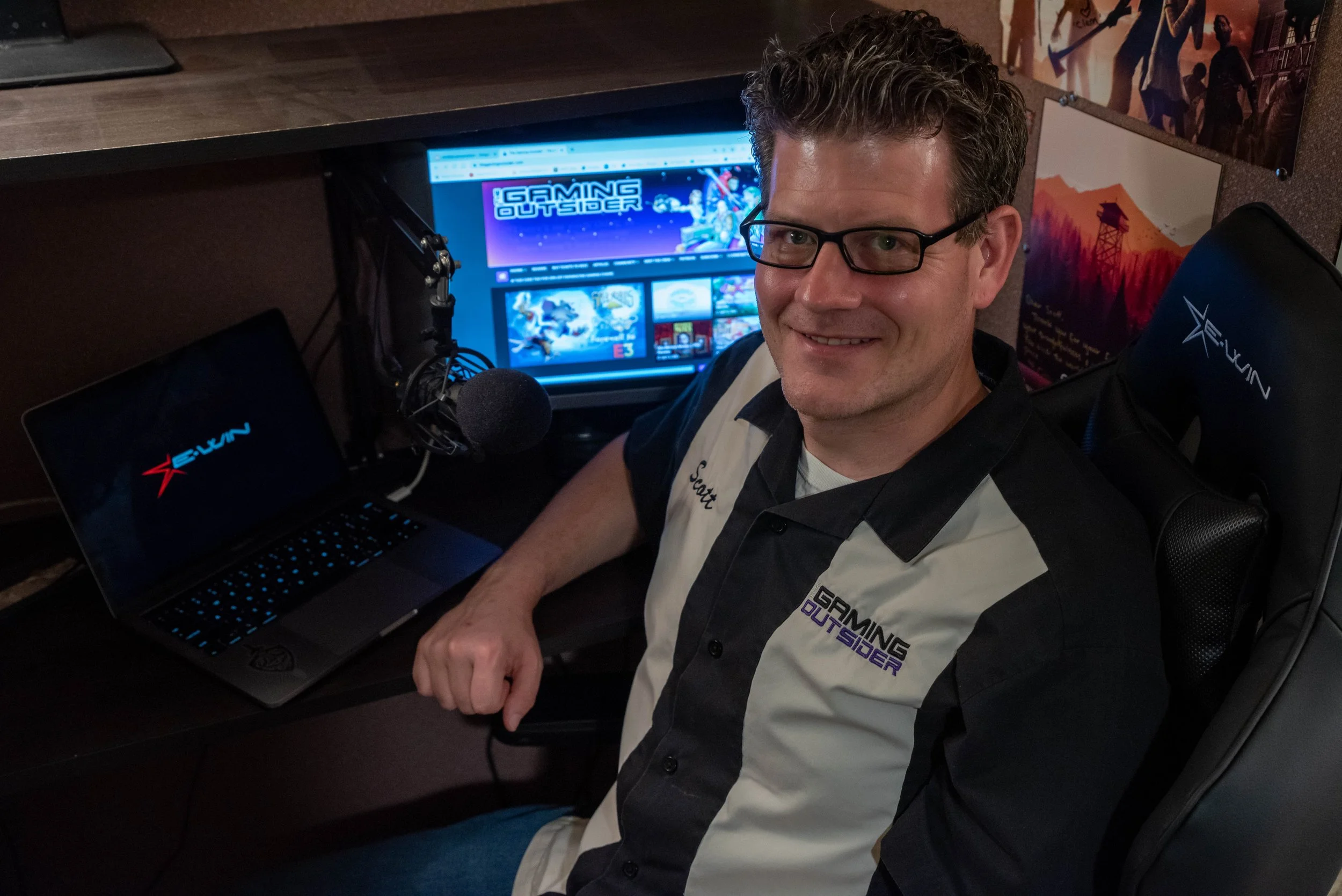 Smiling man with glasses sitting at a desk with gaming equipment, including a monitor displaying a gaming website, a laptop, a microphone, and posters on the wall.