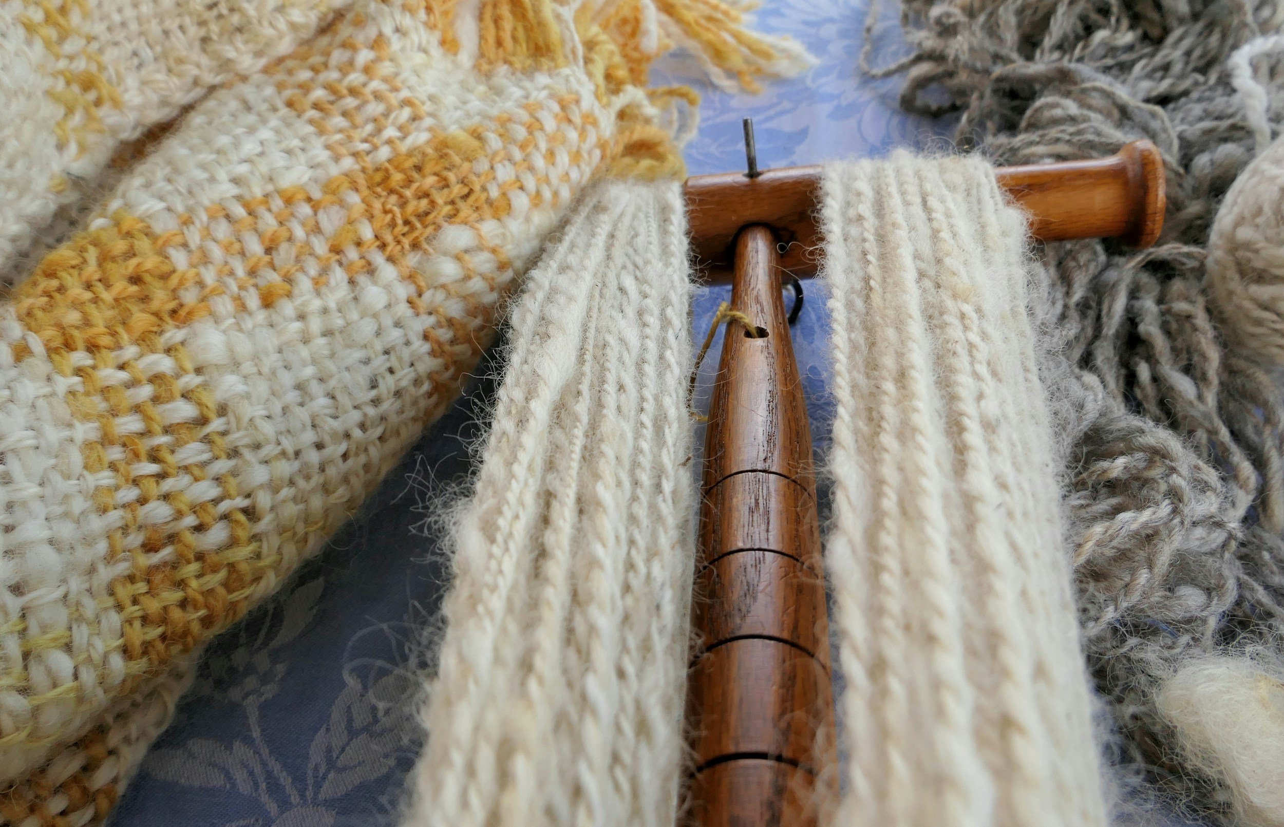 Close-up of a wooden weaving loom with several threads of yarn in cream and gray colors, surrounded by completed woven fabric pieces in cream with yellow accents.
