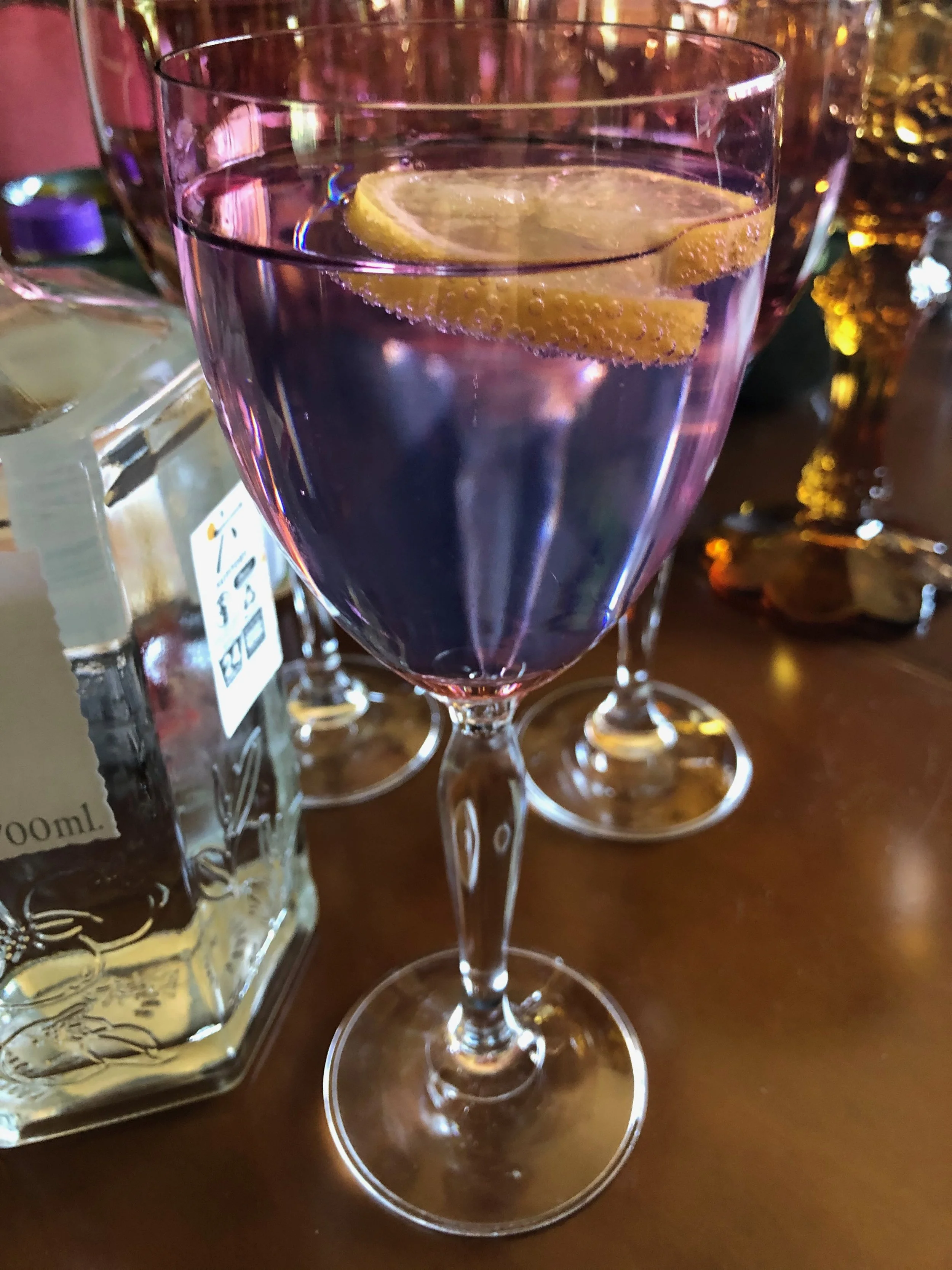 A close-up of a glass of purple-colored drink with a lemon slice inside. In the background, there are other glasses and bottles on a wooden surface.