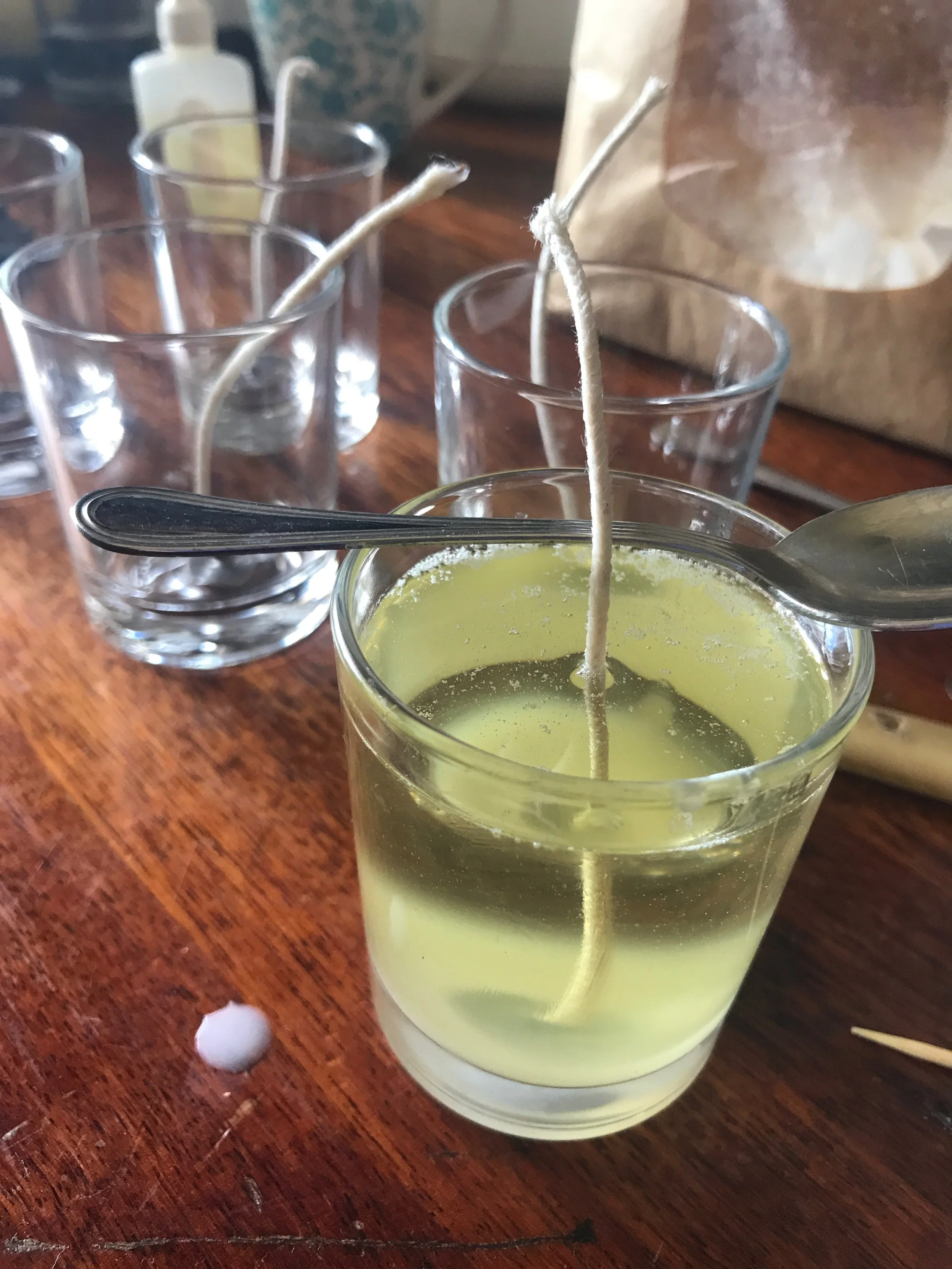 A glass of greenish-yellow liquid with a chopstick and spoon inside on a wooden table, with three empty glasses with chopsticks in the background, and a golf ball and toothpick on the table.