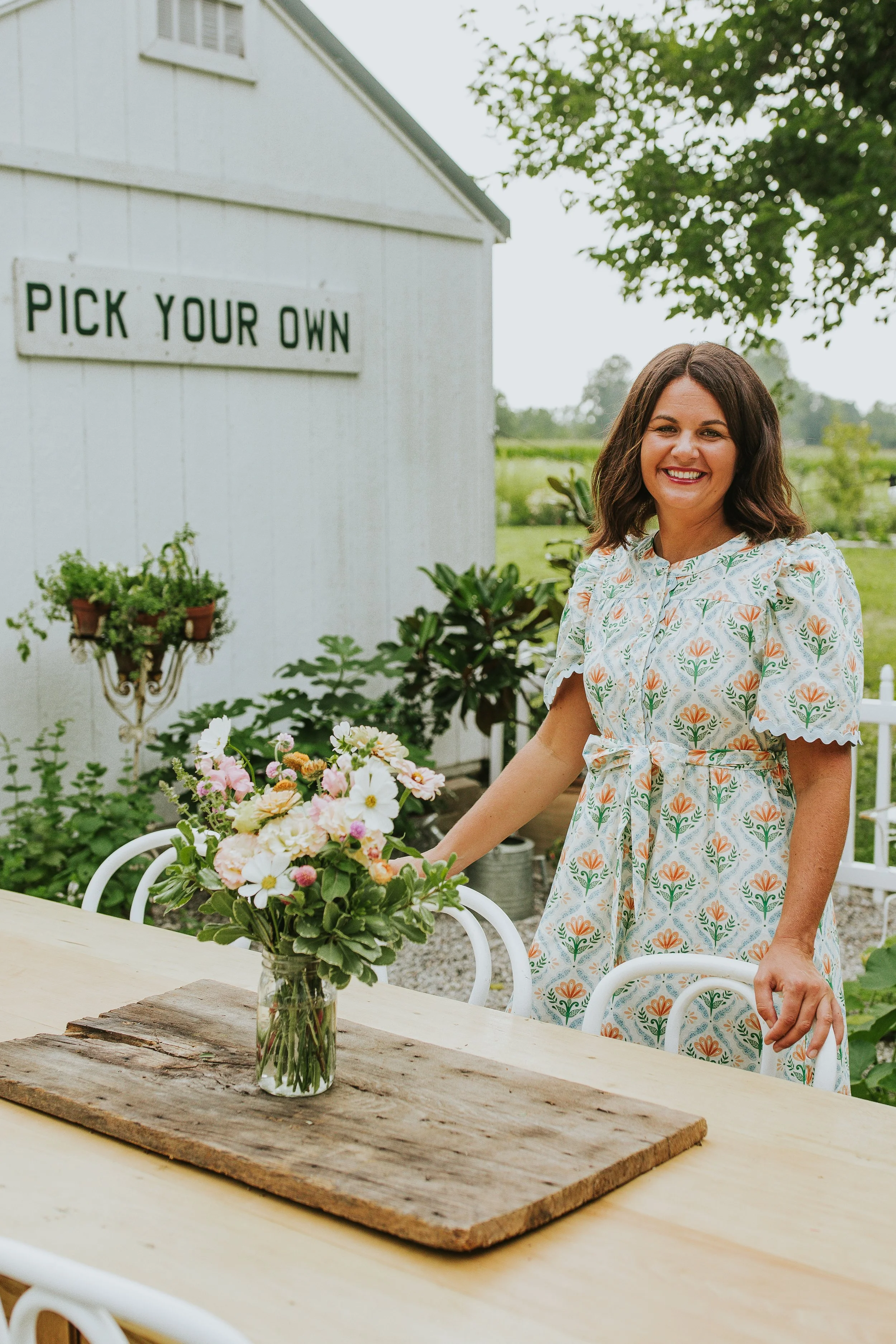 A woman in a floral dress standing outdoors near a table with a vase of colorful flowers. Behind her is a white building with a sign that reads "PICK YOUR OWN" and some potted plants. She is smiling and holding a chair.