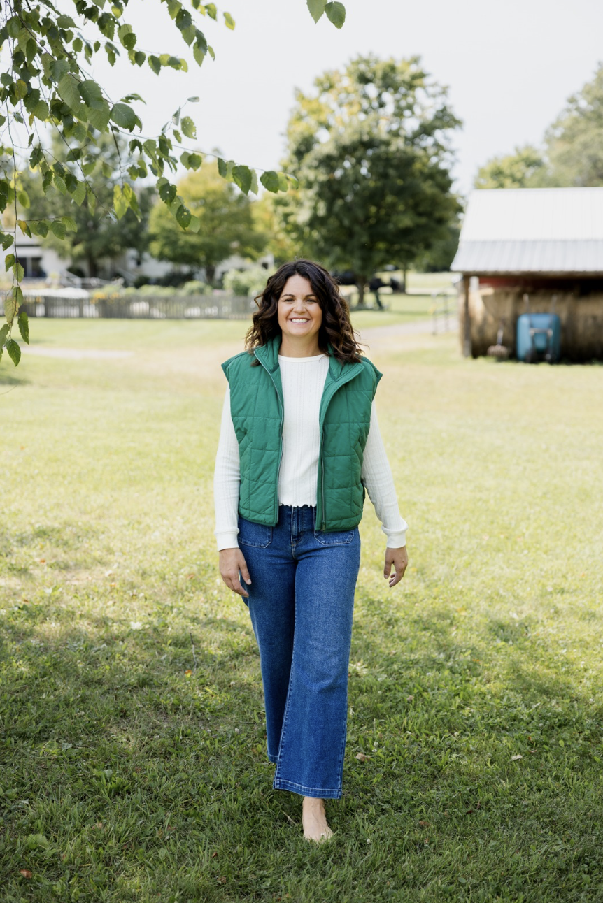 A woman smiling outside on a grassy field with trees and a wooden building in the background.