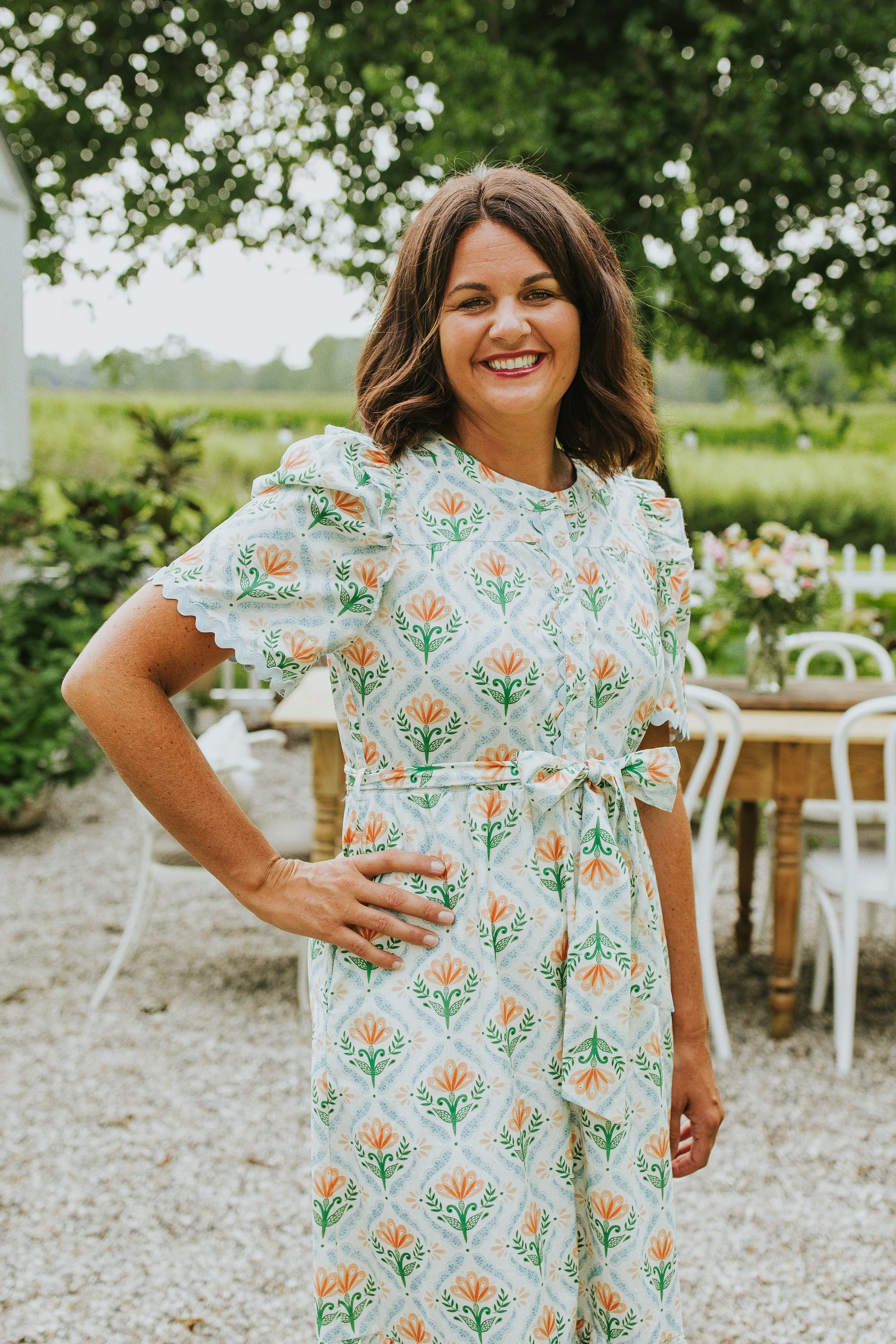 A woman standing outdoors in a garden or park, smiling and wearing a floral patterned dress with short sleeves.