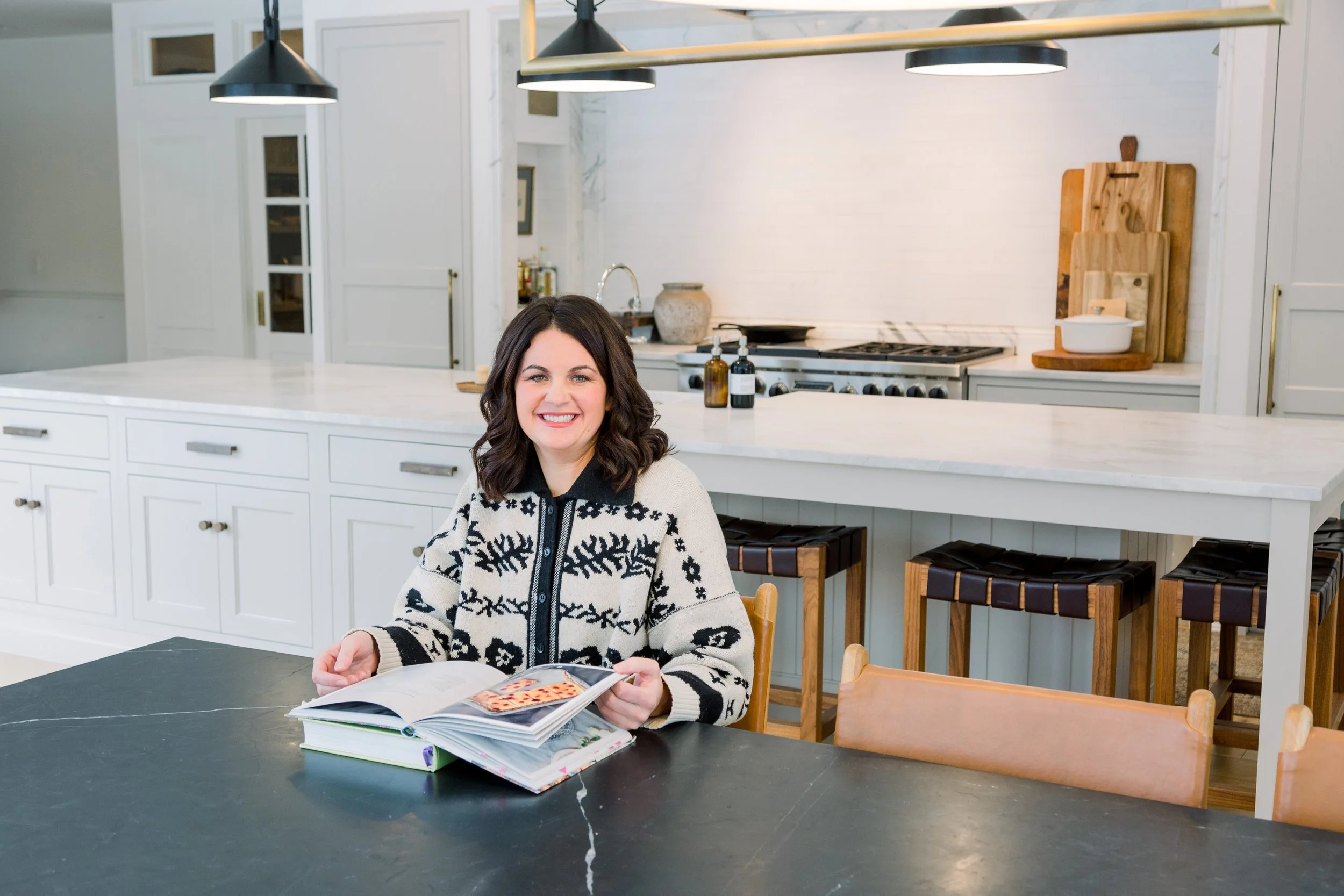 A woman with dark hair, smiling, sitting at a kitchen island reading a book with colorful pages, in a modern kitchen with white cabinets and wooden accents.