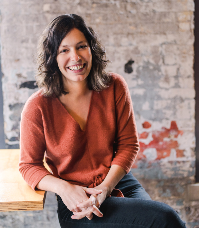 A woman with dark, wavy hair smiling and sitting on a chair against a rustic brick wall, wearing a rust-colored sweater.