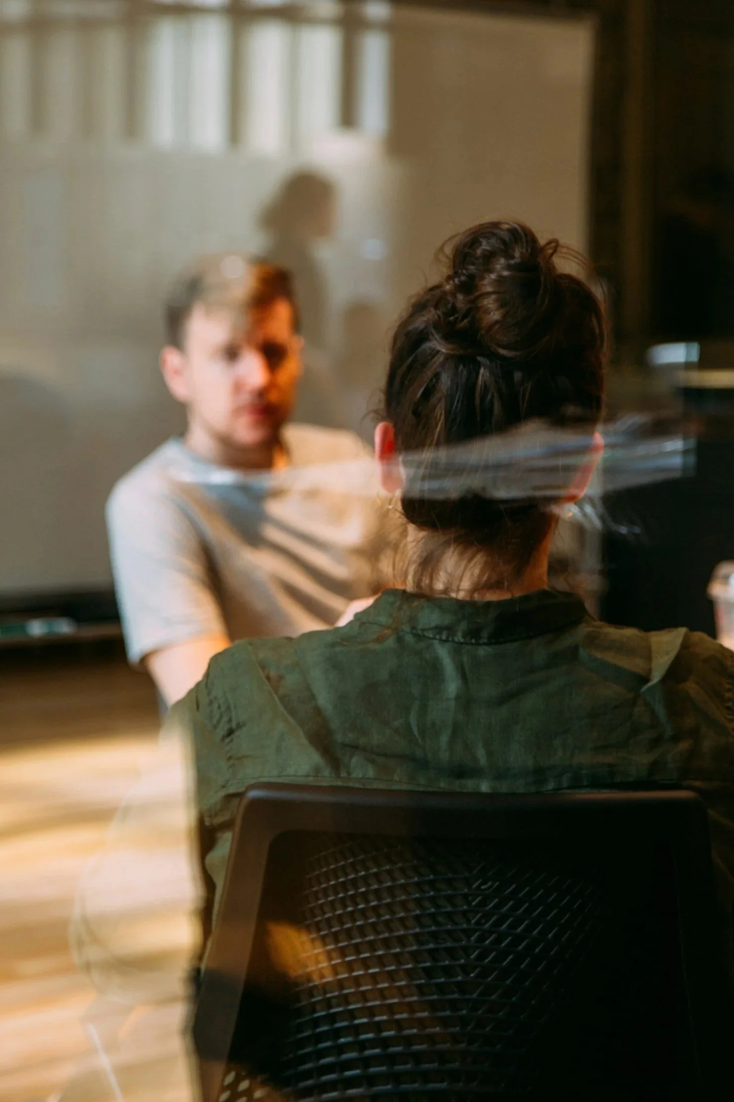 The image shows a woman sitting with her back to the camera, facing a man who is slightly blurry and looking towards her. They are indoors, with a curtain and window in the background.