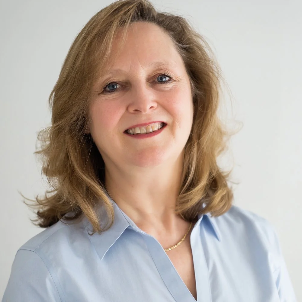 A woman with shoulder-length light brown hair and blue eyes, smiling while wearing a light blue collared shirt and a gold necklace.