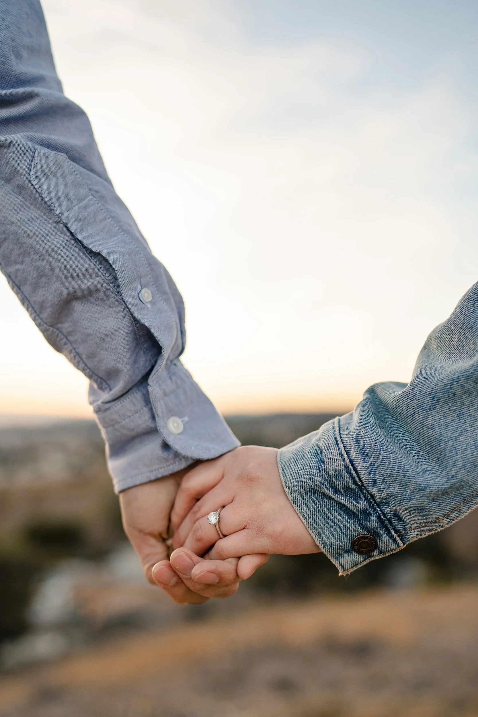 A close-up of two people holding hands, one person wearing a gray button-up shirt and the other wearing a denim jacket, with a blurred outdoor background and a cloudy sky.