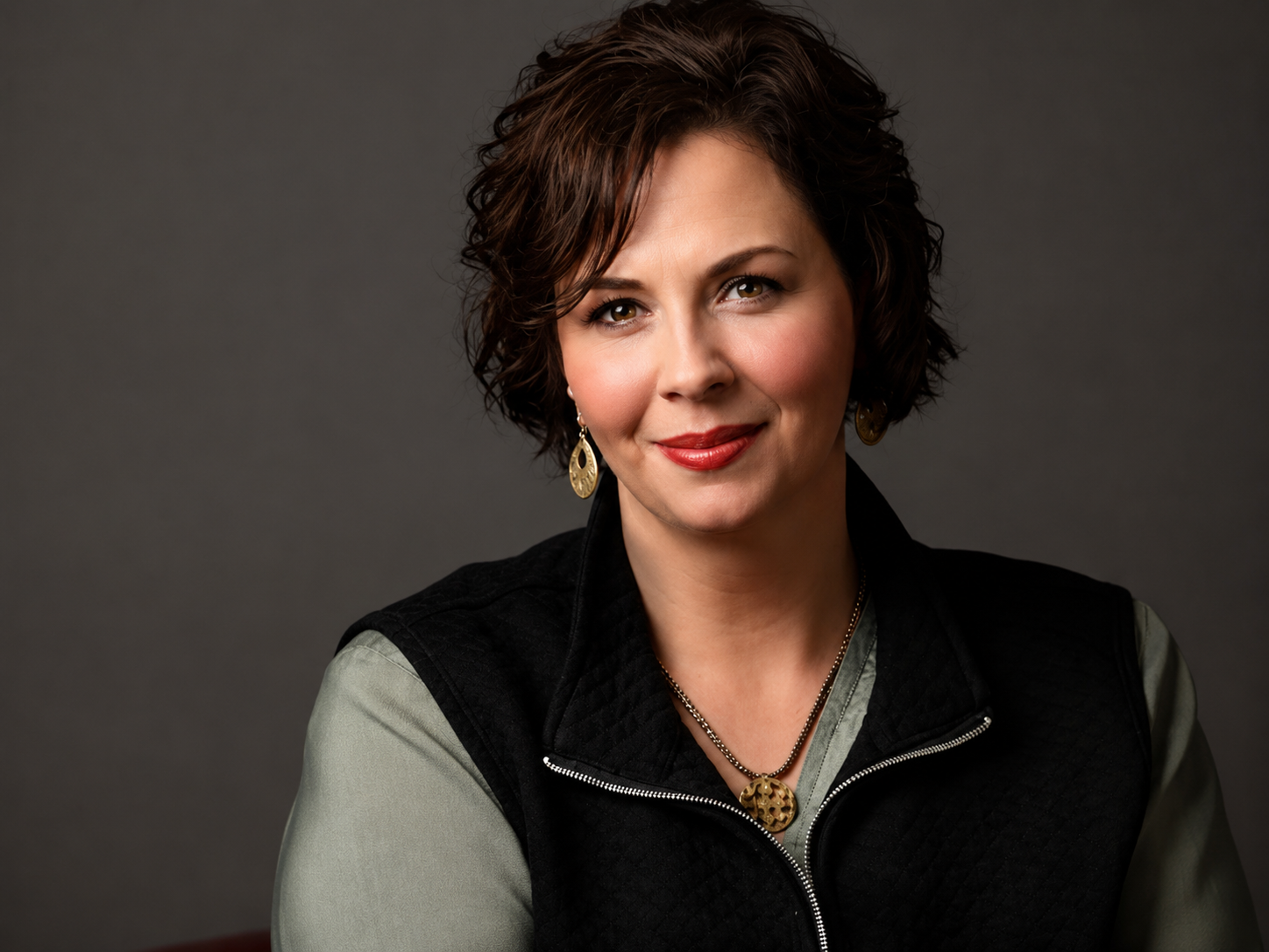 Portrait of a woman with short wavy brown hair, wearing red lipstick, gold earrings, a necklace with a gold pendant, a light green shirt, and a black vest, against a dark gray background.