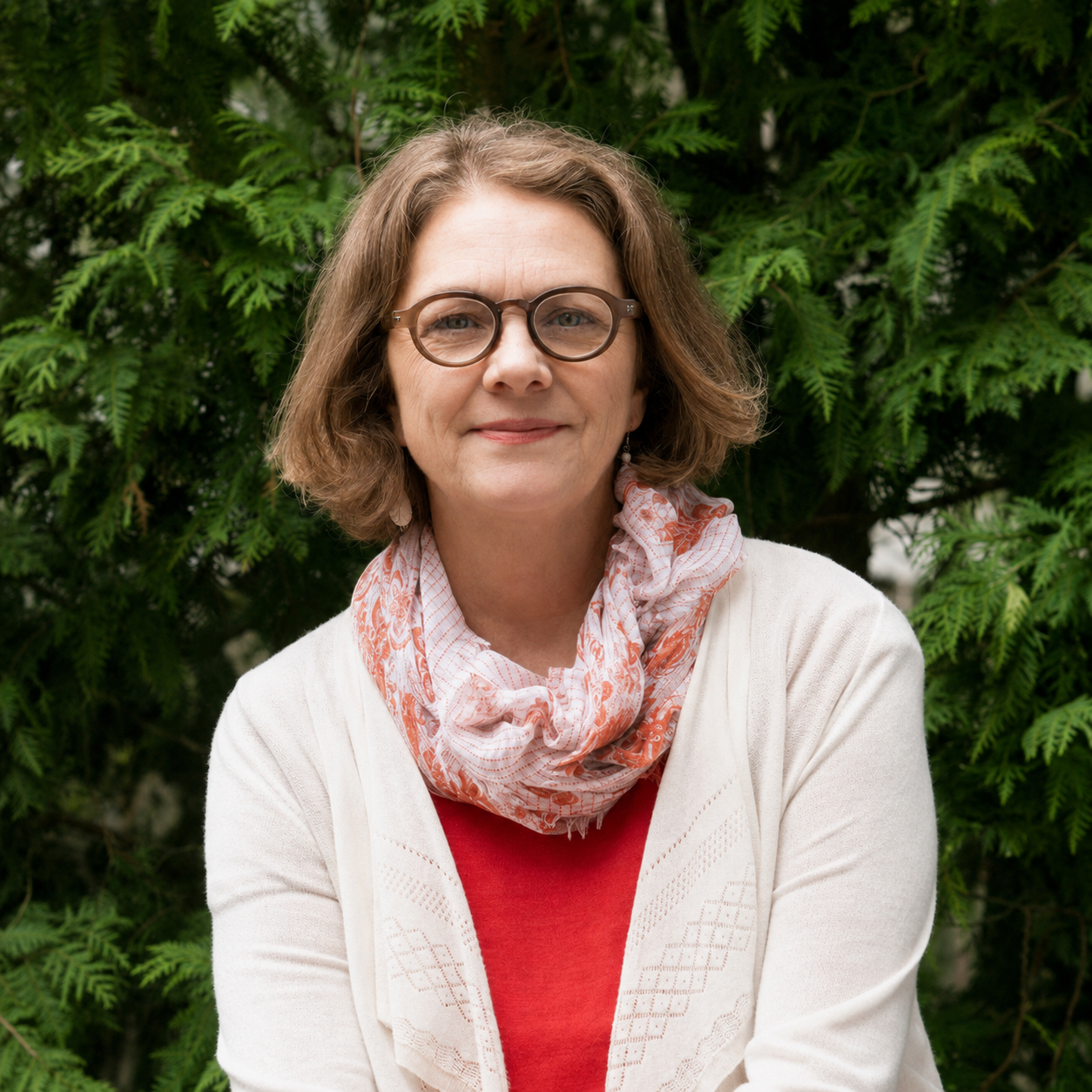 A woman with shoulder-length brown hair, glasses, and light skin, smiling outdoors against green foliage; wearing a white cardigan over a red shirt and a pink patterned scarf.