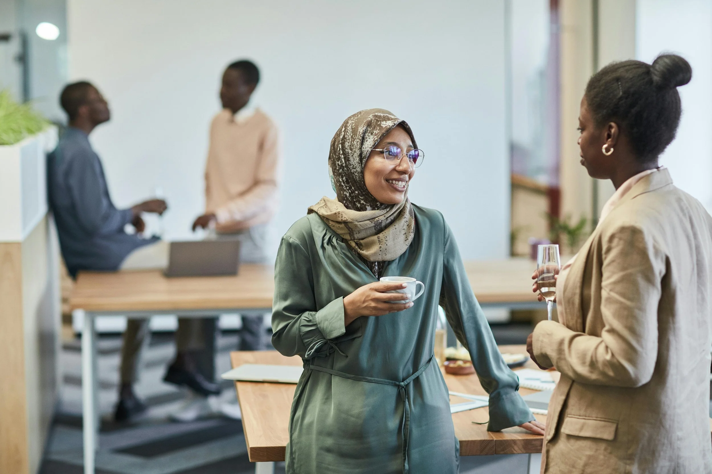 Two women are engaged in conversation in an office or conference room. One woman is wearing glasses and a headscarf, holding a cup, and the other woman has her hair styled in a bun. In the background, two other women are talking near a counter.