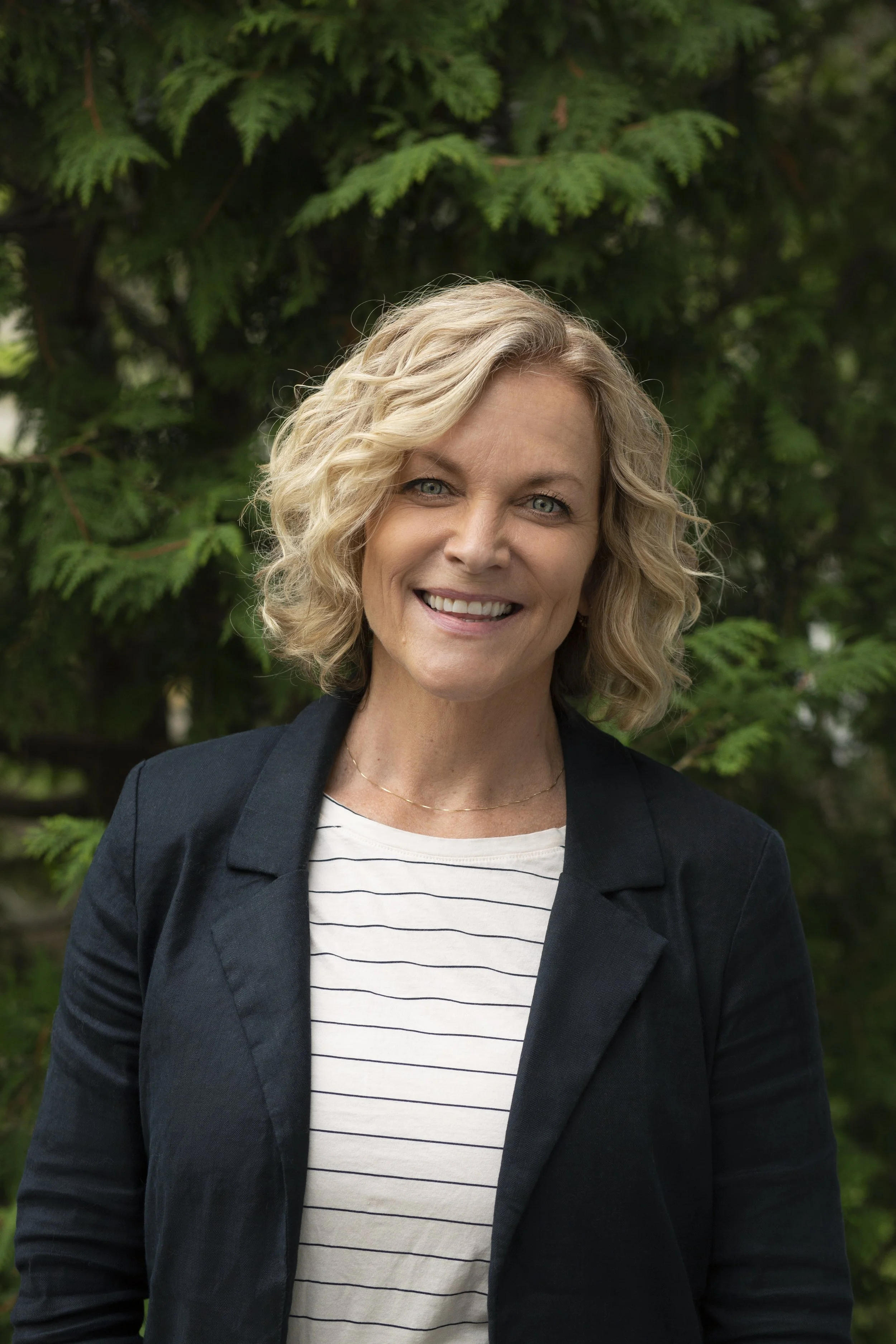 A woman with shoulder-length wavy blonde hair wearing a dark blazer and a white striped shirt, standing outdoors in front of green foliage, smiling at the camera.
