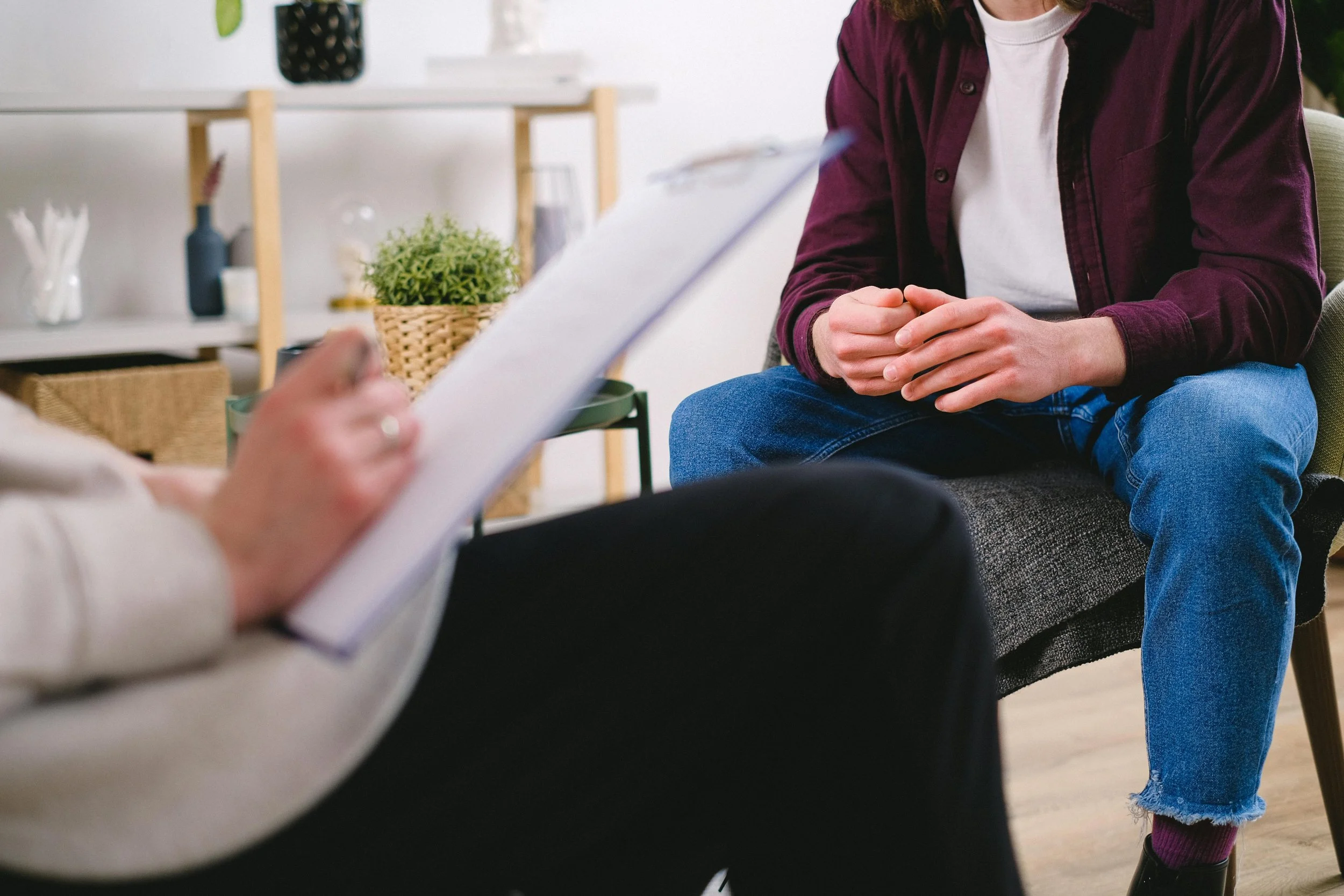 A person wearing a maroon shirt and ripped jeans sitting with hands clasped during a conversation, with another person holding a clipboard partially visible in a casual indoor setting.