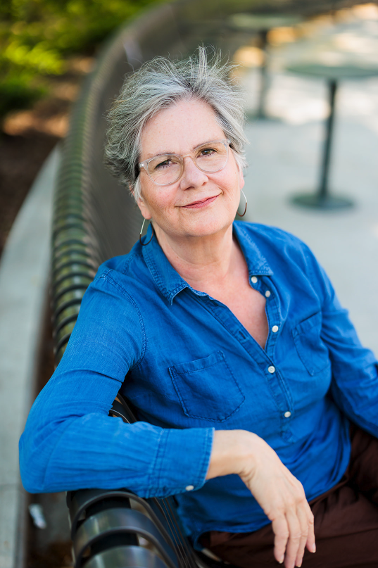 An older woman with short gray hair, glasses, and hoop earrings smiling while sitting on a park bench, wearing a blue button-up shirt.