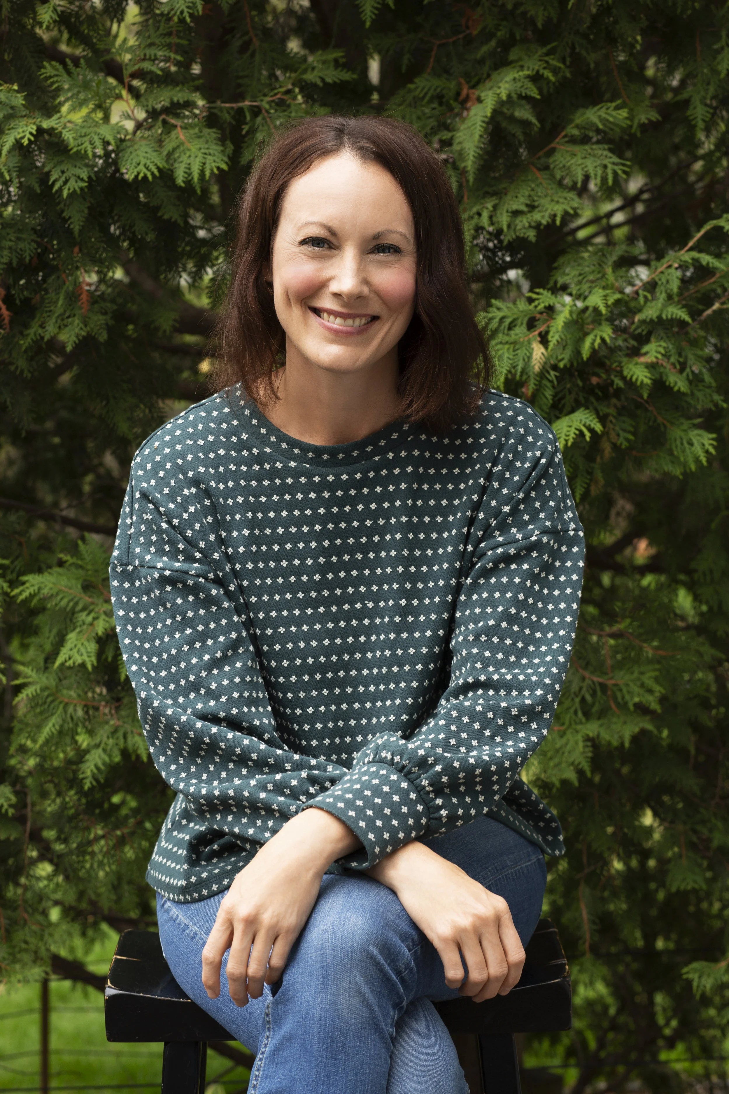 A woman with brown hair smiling while sitting outdoors in front of green foliage, wearing a dark green sweater with white polka dots and blue jeans.